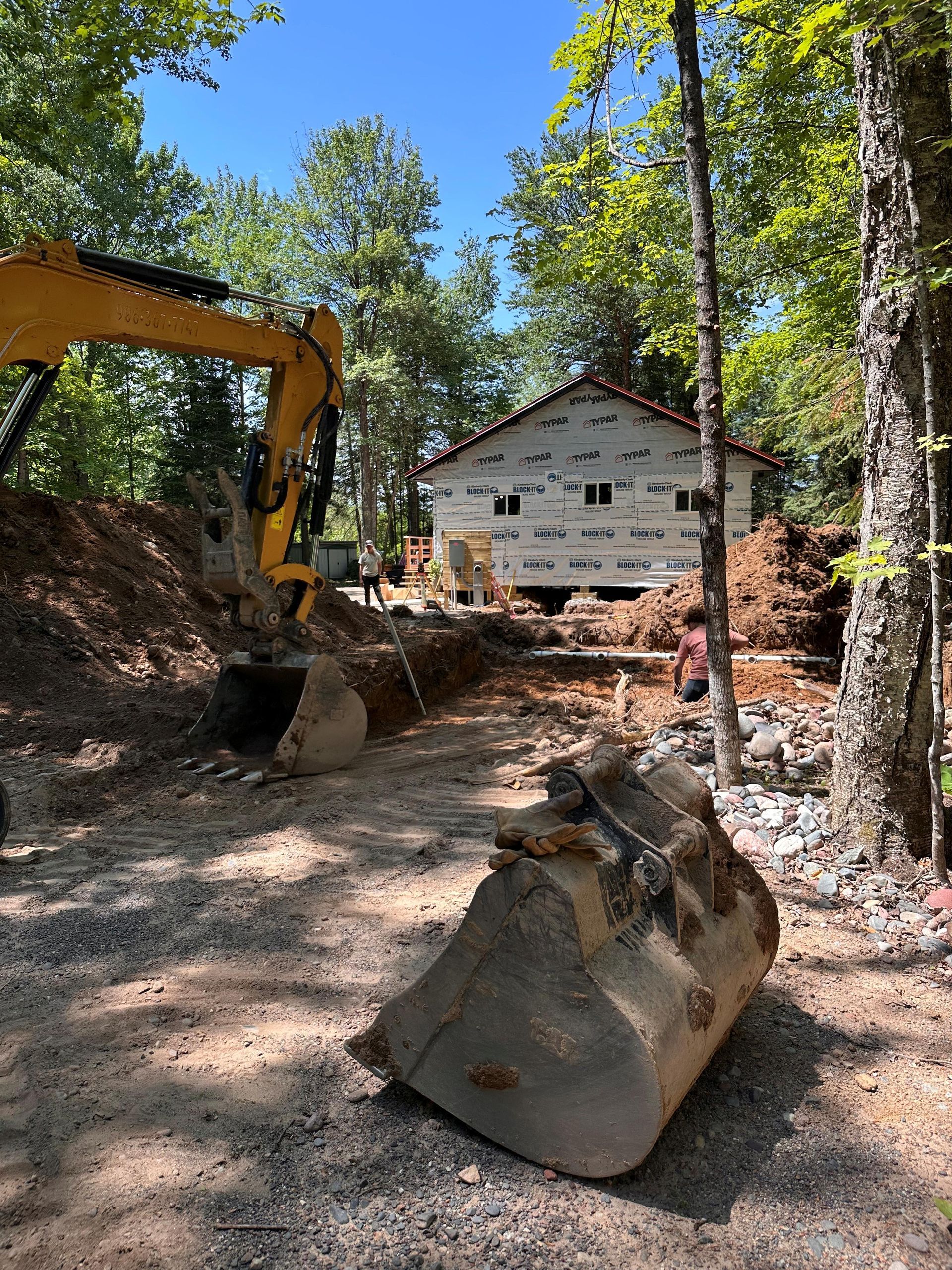 A yellow excavator works in a dirt clearing near a house under construction with white exterior sheeting. A yellow excavator works in a dirt clearing near a house under construction with white exterior sheeting.