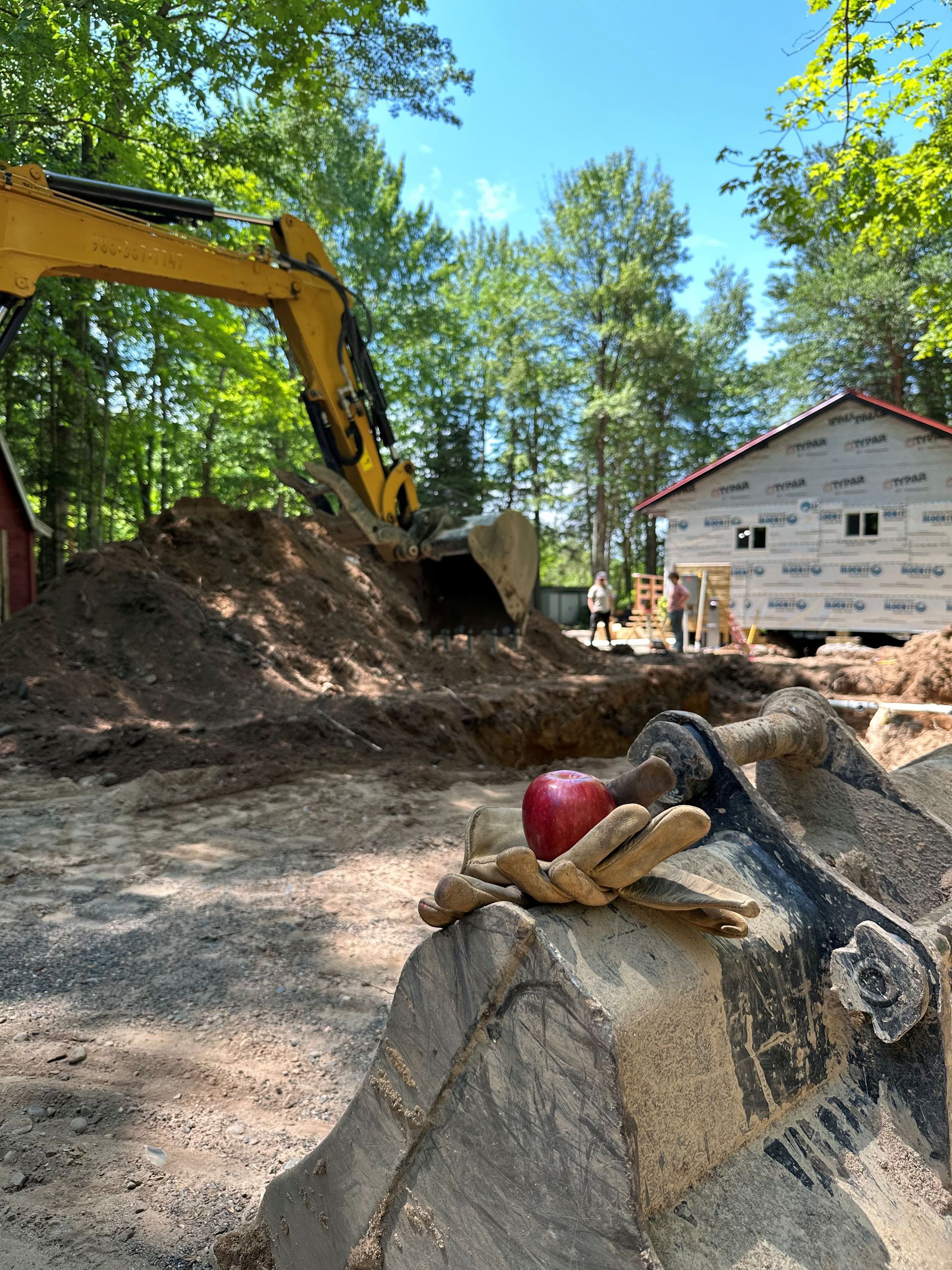 An apple rests on a tan work glove placed atop a yellow excavator bucket at a construction site with a house under way. An apple rests on a tan work glove placed atop a yellow excavator bucket at a construction site with a house under way.