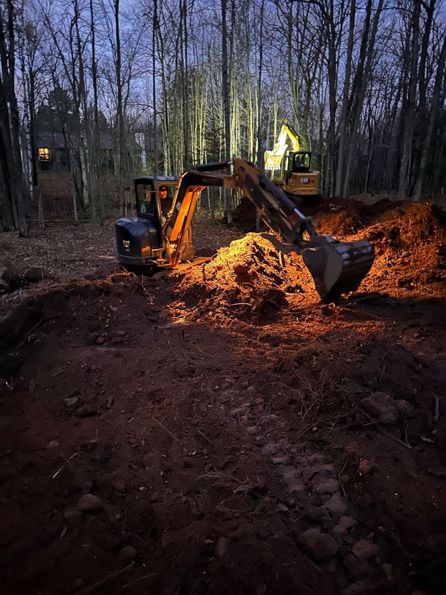 A yellow excavator at night works in a wooded area, scooping a large pile of dirt with its illuminated bucket.