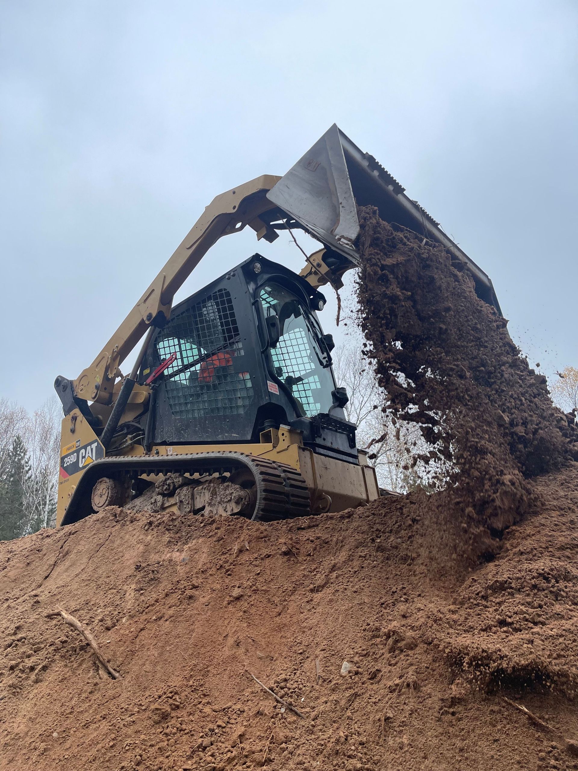 A yellow Caterpillar compact track loader on a dirt pile, dumping a load of soil from its raised bucket.