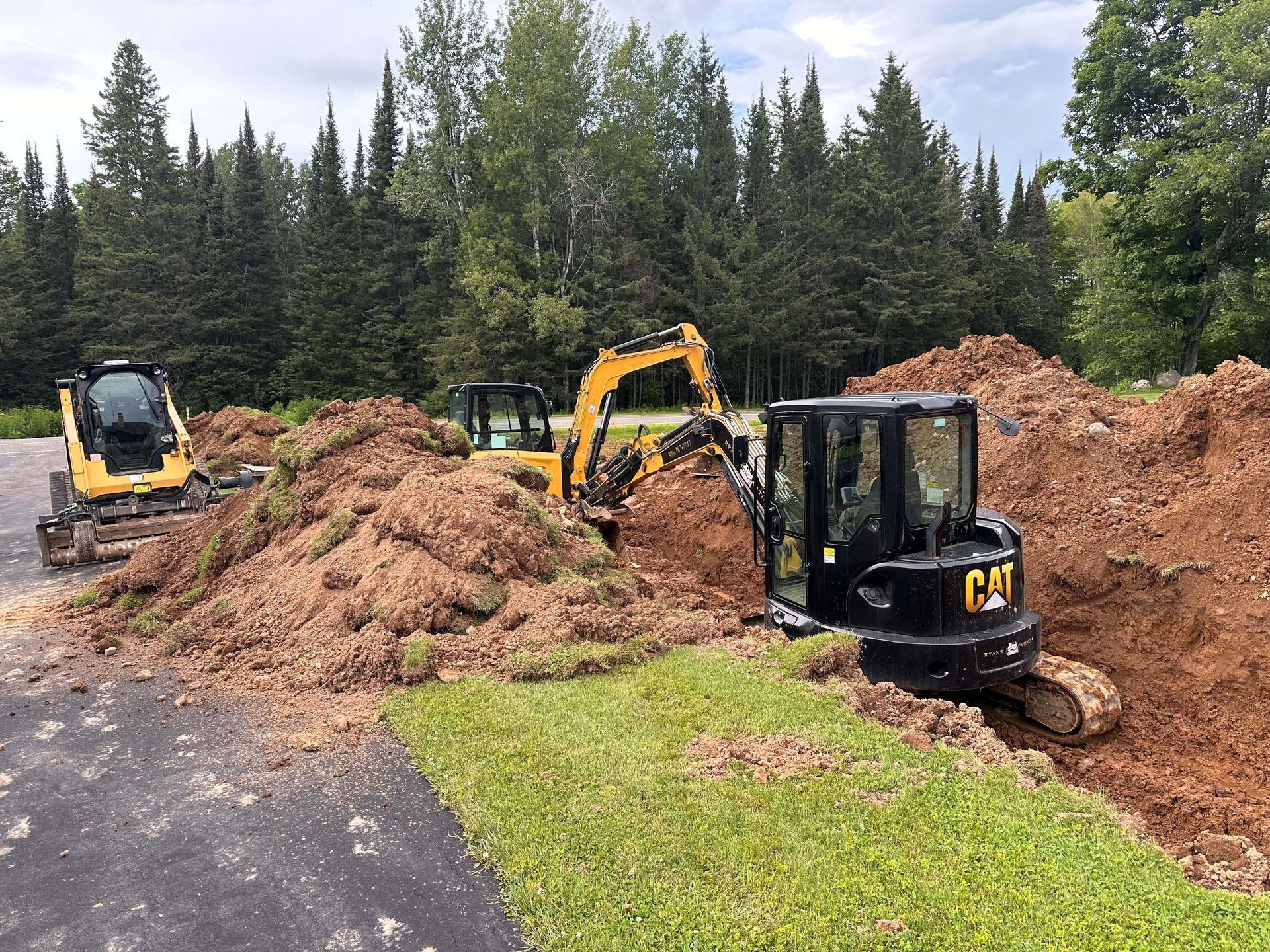 Two yellow and black Caterpillar excavators digging in piles of dirt near a wooded area and a paved driveway. Two yellow and black Caterpillar excavators digging in piles of dirt near a wooded area and a paved driveway.