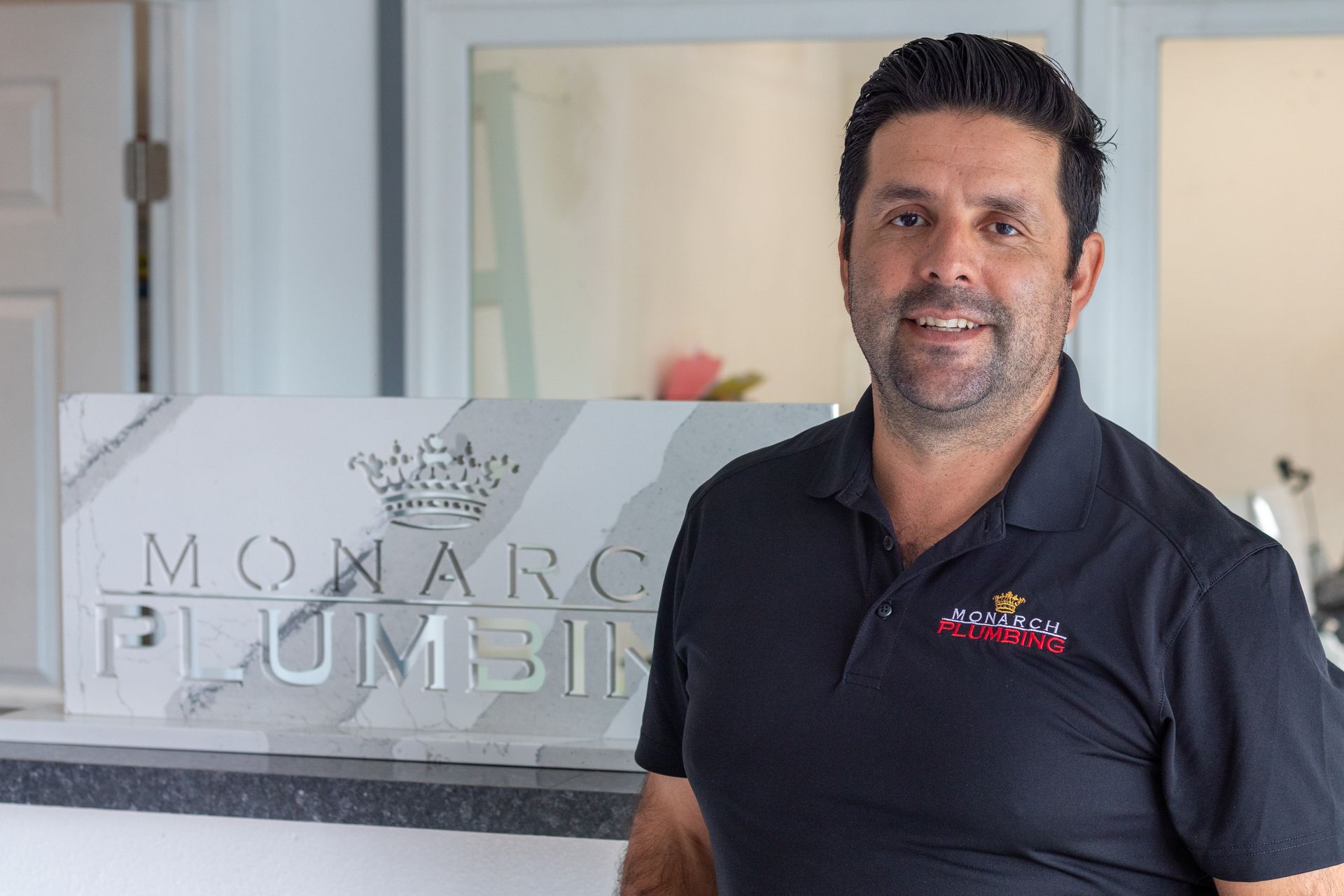 Jose the CEO of Monarch Plumbing standing beside a frosted glass sign reading “Monarch Plumbing” in an office lobby