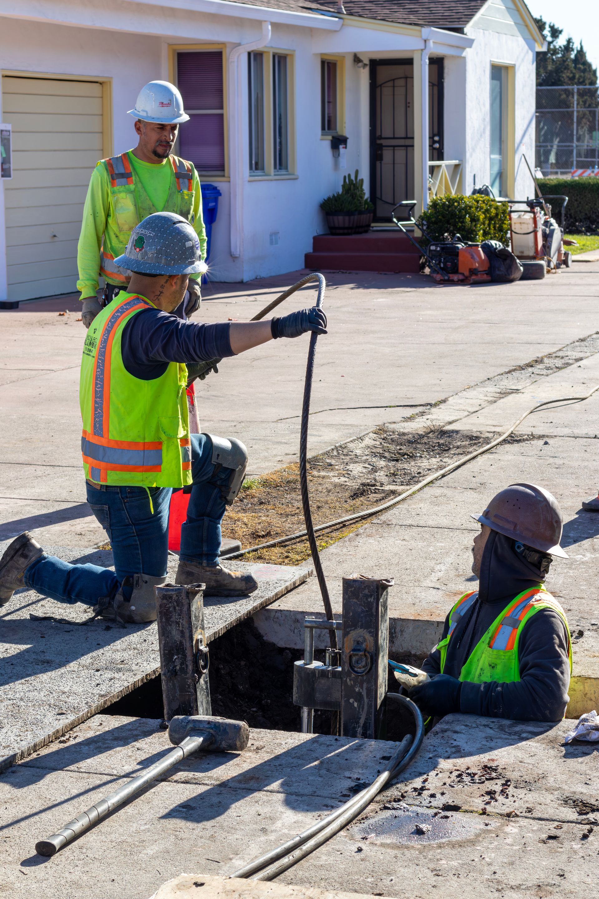 Workers prepping for sewer replacement, with one person using a jackhammer and orange traffic cones nearby.