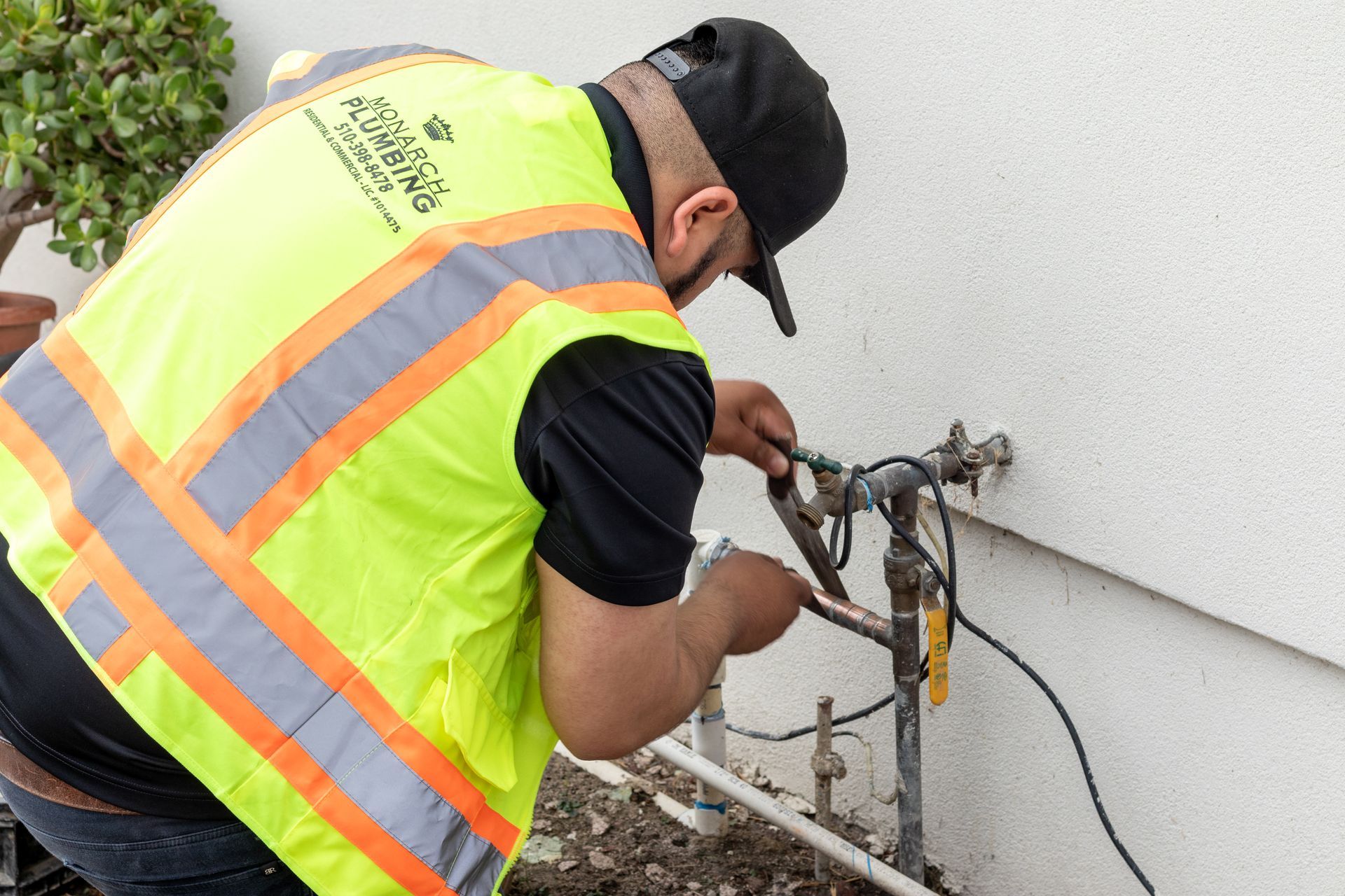 Monarch Plumbing worker in a neon safety vest repairs outdoor utility piping beside a building.