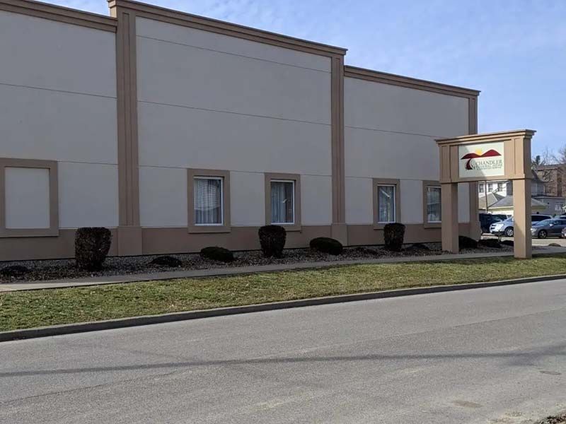 A low, beige building with a sign, set on a curb beside a road. The sky is blue.
