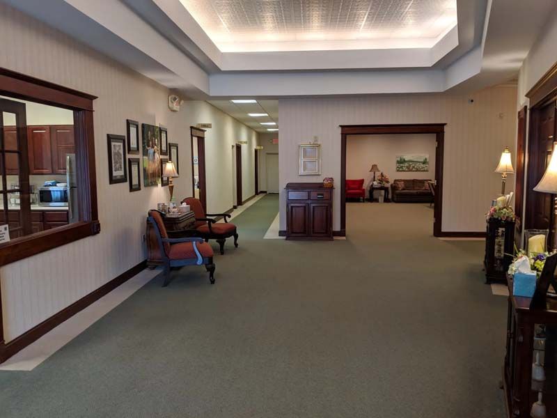 Hallway with chairs, framed photos, and an open doorway to a sitting area with a person.