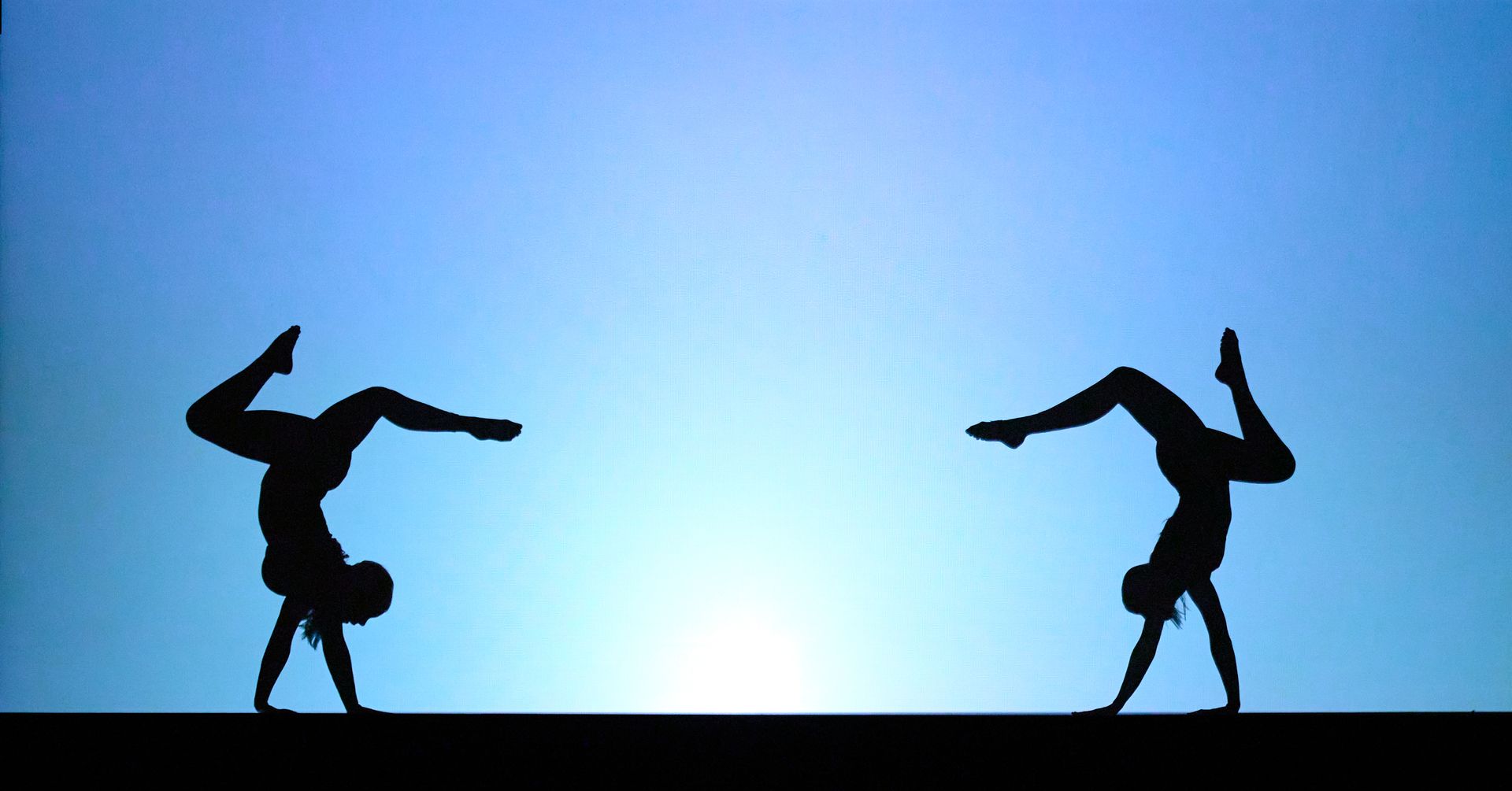 Two black dancer silhouettes balancing in mirrored poses against a blue gradient background.