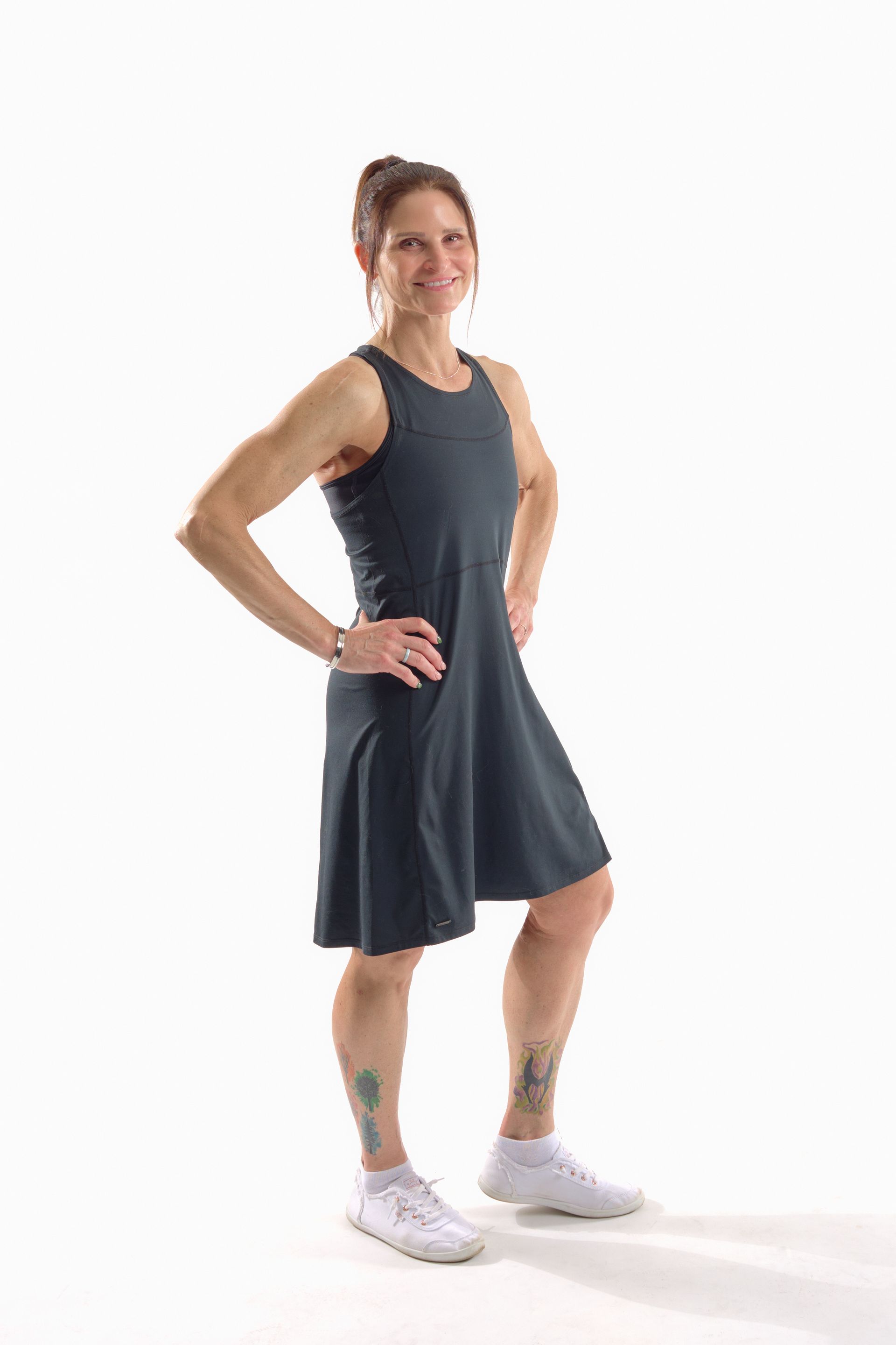 Woman in dark athletic dress and white sneakers standing with hands on hips against a white background