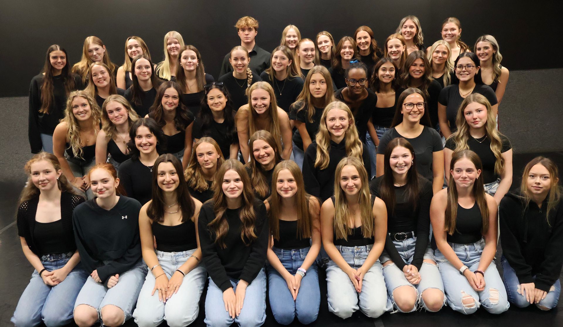 Large group posing for a team photo, wearing coordinated black tops and light jeans, seated and standing in rows.