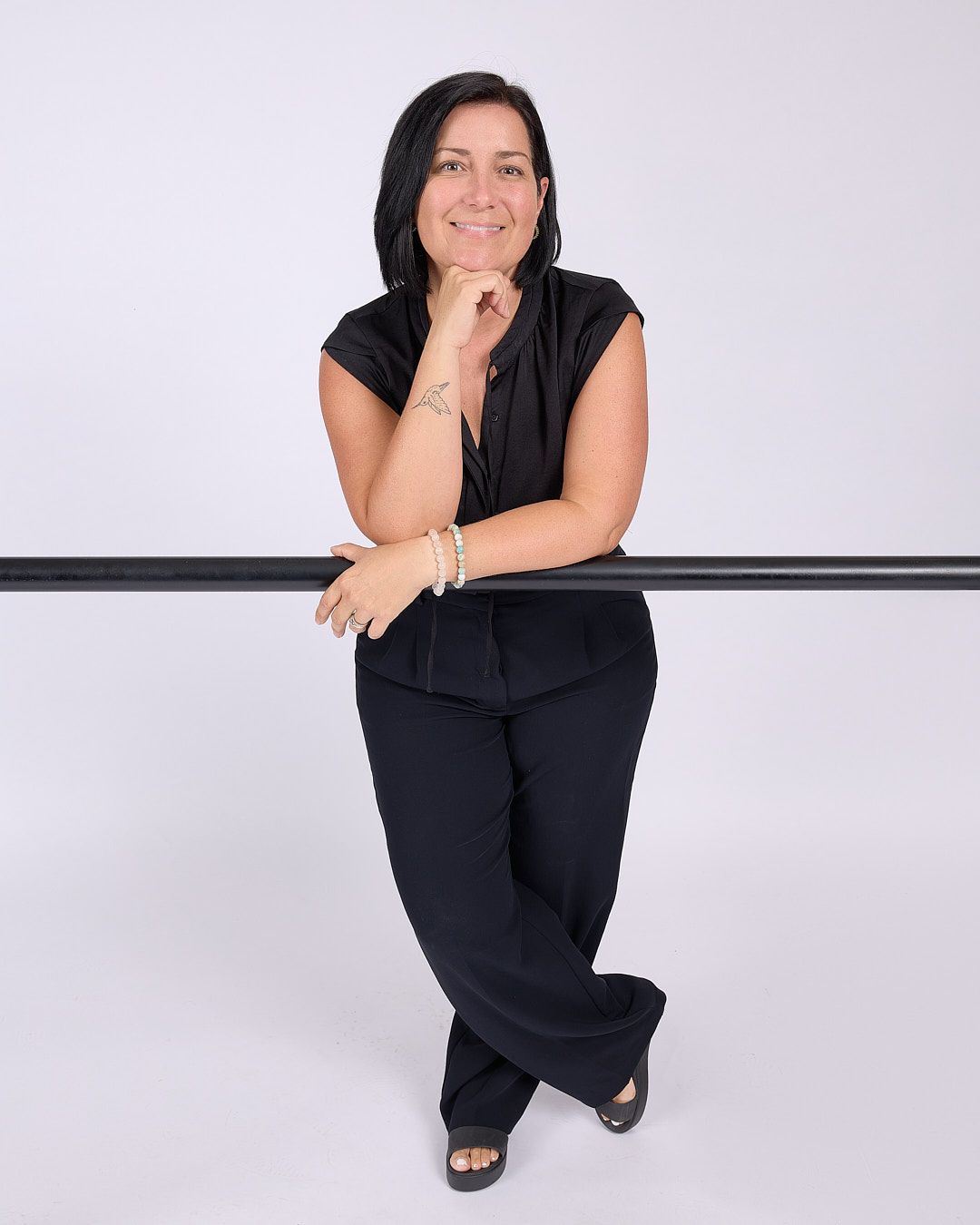 Person in black outfit leaning on a rail, smiling against a light studio background