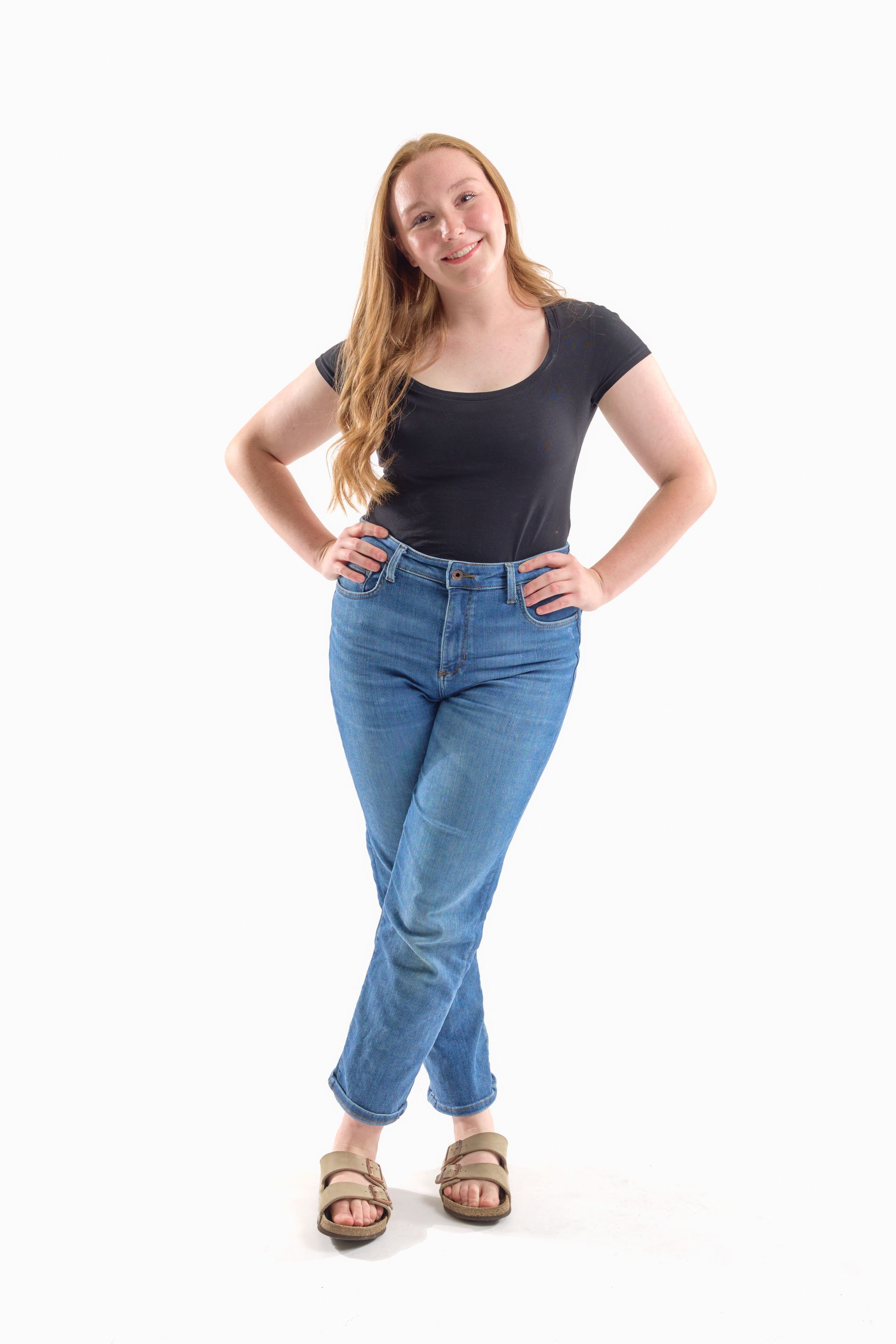Woman standing with hands on hips, wearing a black top, blue jeans, and sandals against a white background