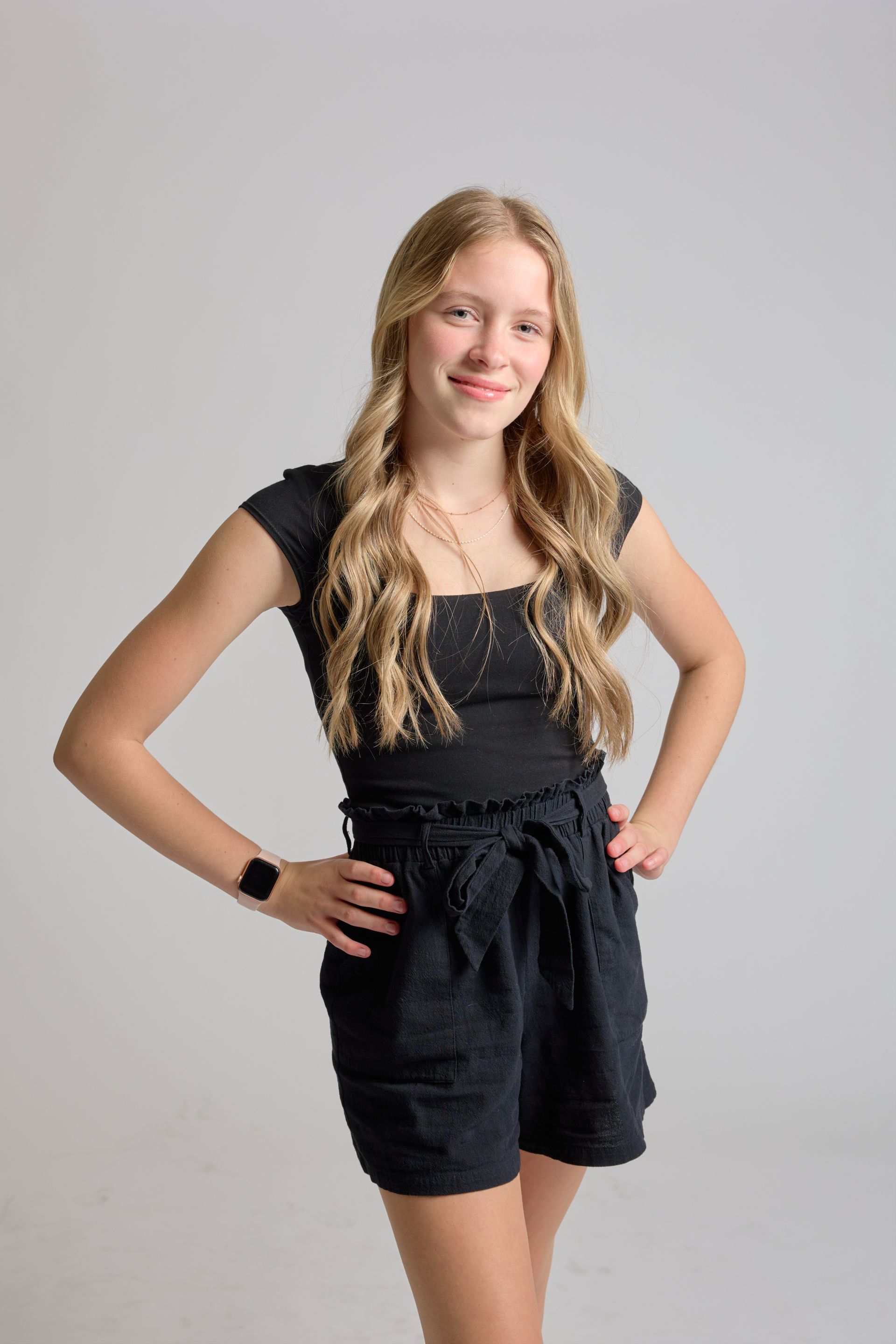 Person in a black romper and smartwatch standing with hands on hips against a gray background