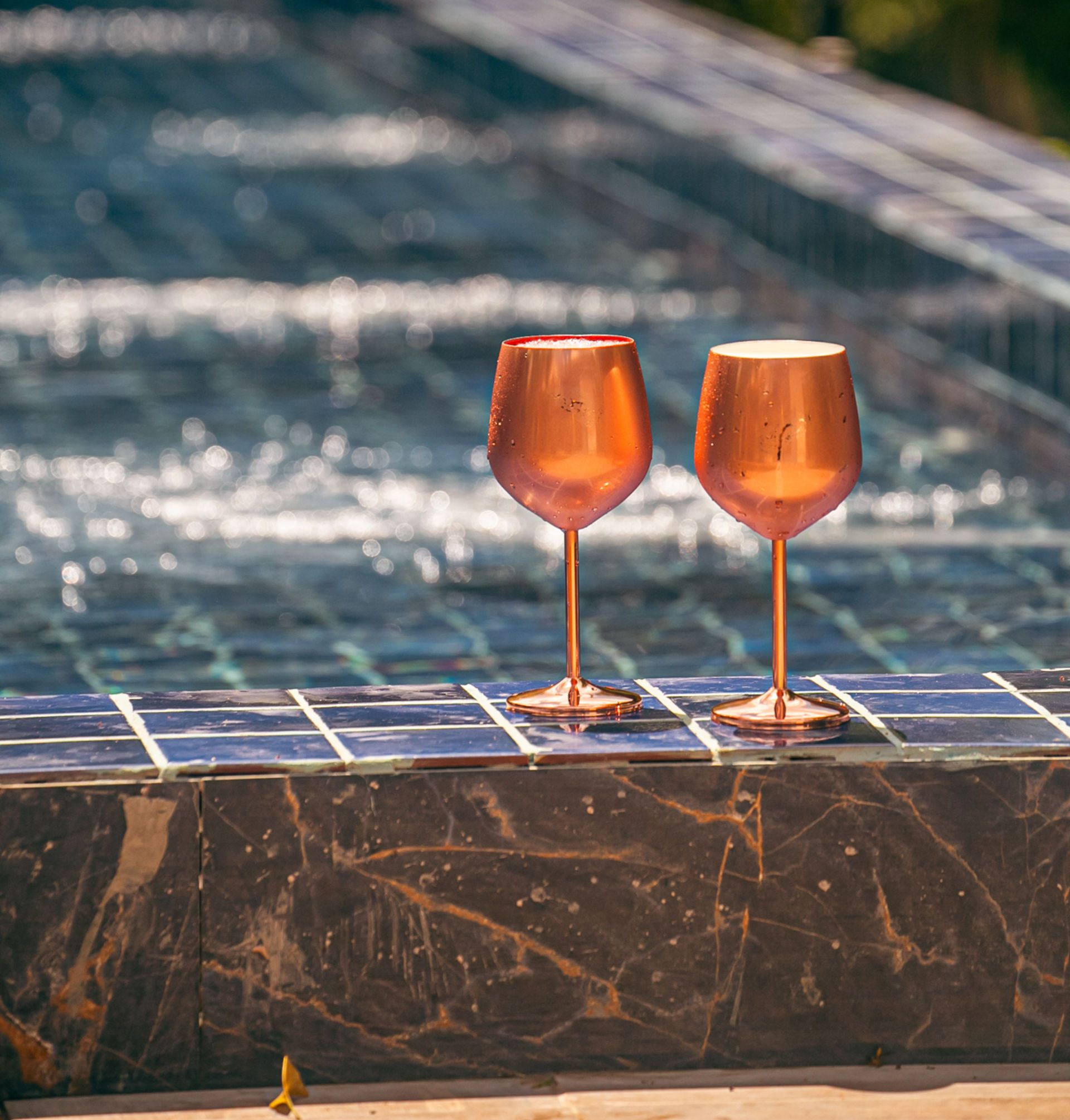 Two copper-colored wine glasses sit poolside near a dark marble ledge, water in the background.