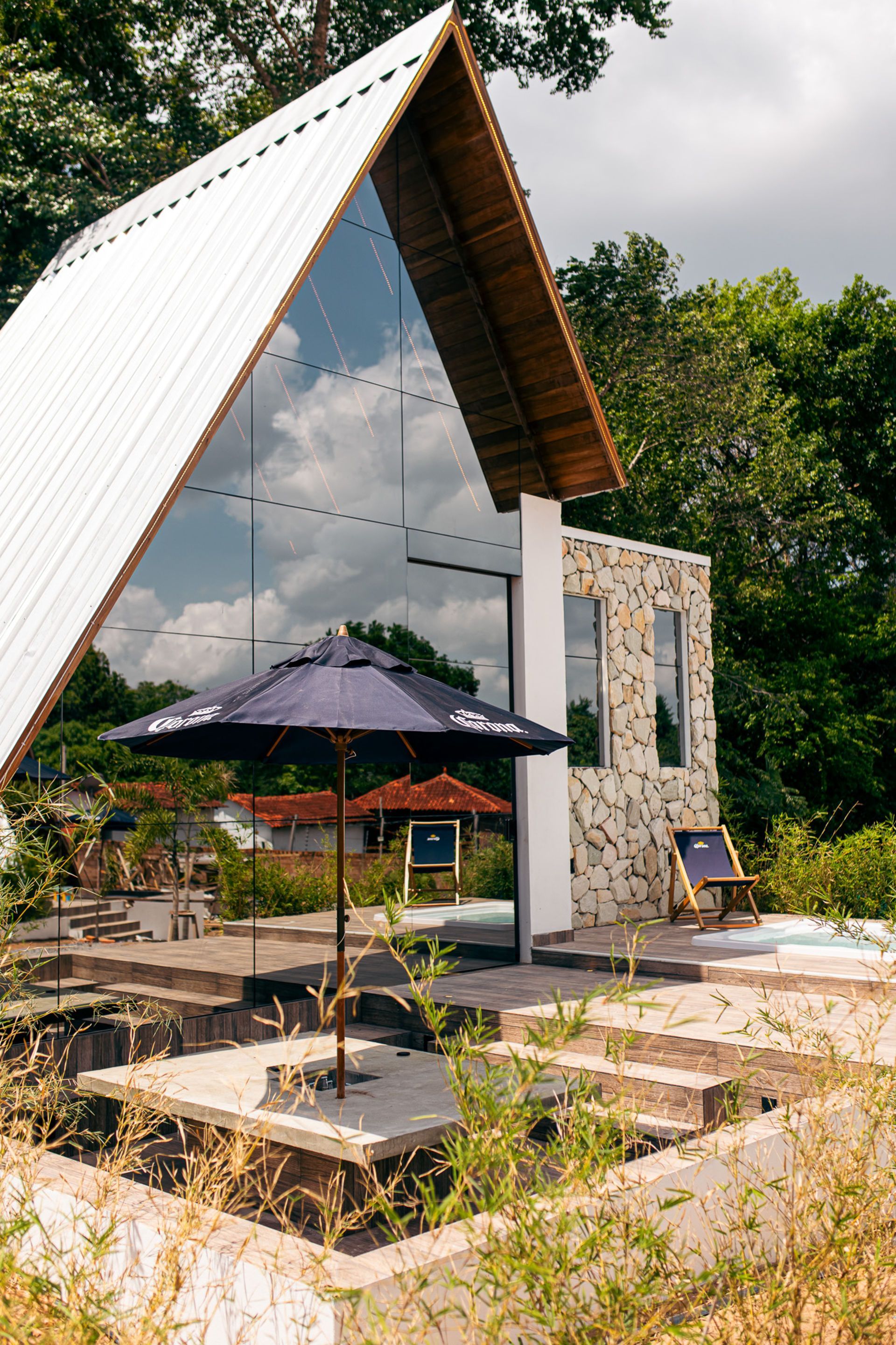 Modern cabin with glass wall reflecting sky, umbrella-covered patio, stone facade, and surrounding greenery.