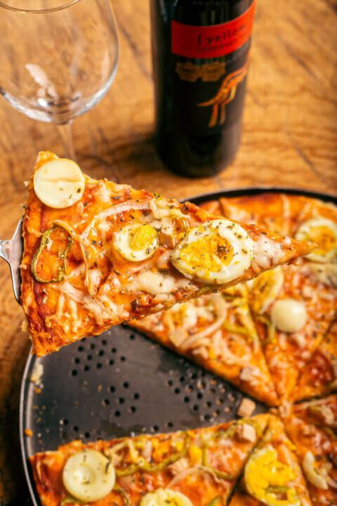Pizza slice topped with egg held by a pizza spatula. Red wine bottle and glass on wooden table.
