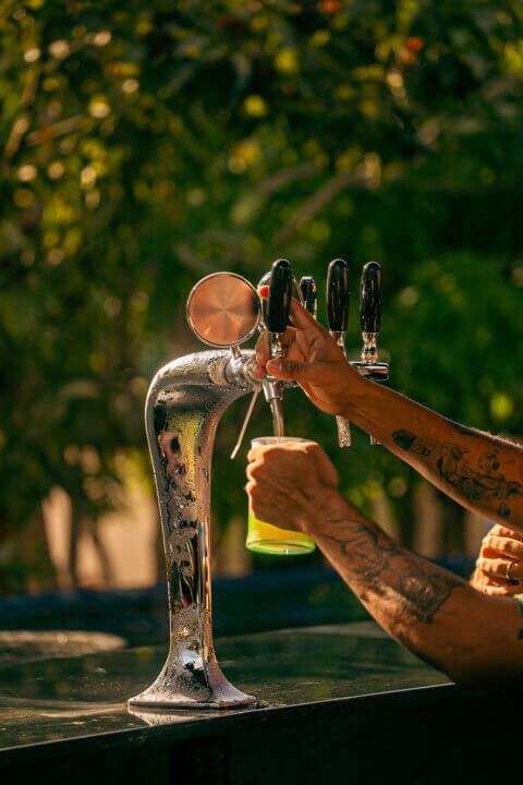 A bartender pouring a draft beer, tattoos visible on their arm, outdoors in the sunlight.