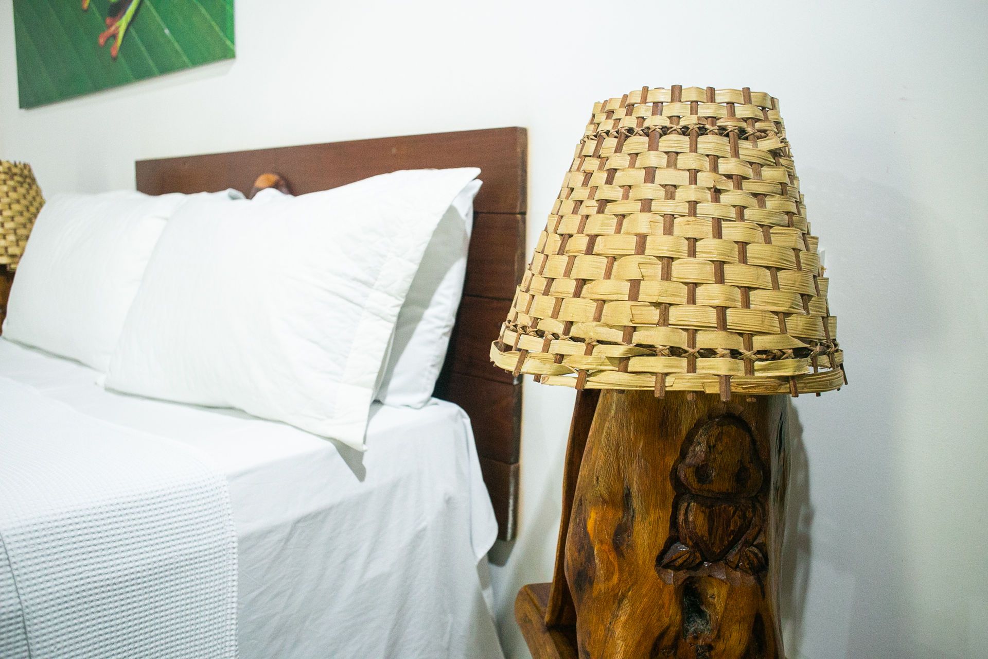 Bedroom with white bedding, a wooden headboard, and a woven lampshade on a carved wooden lamp.