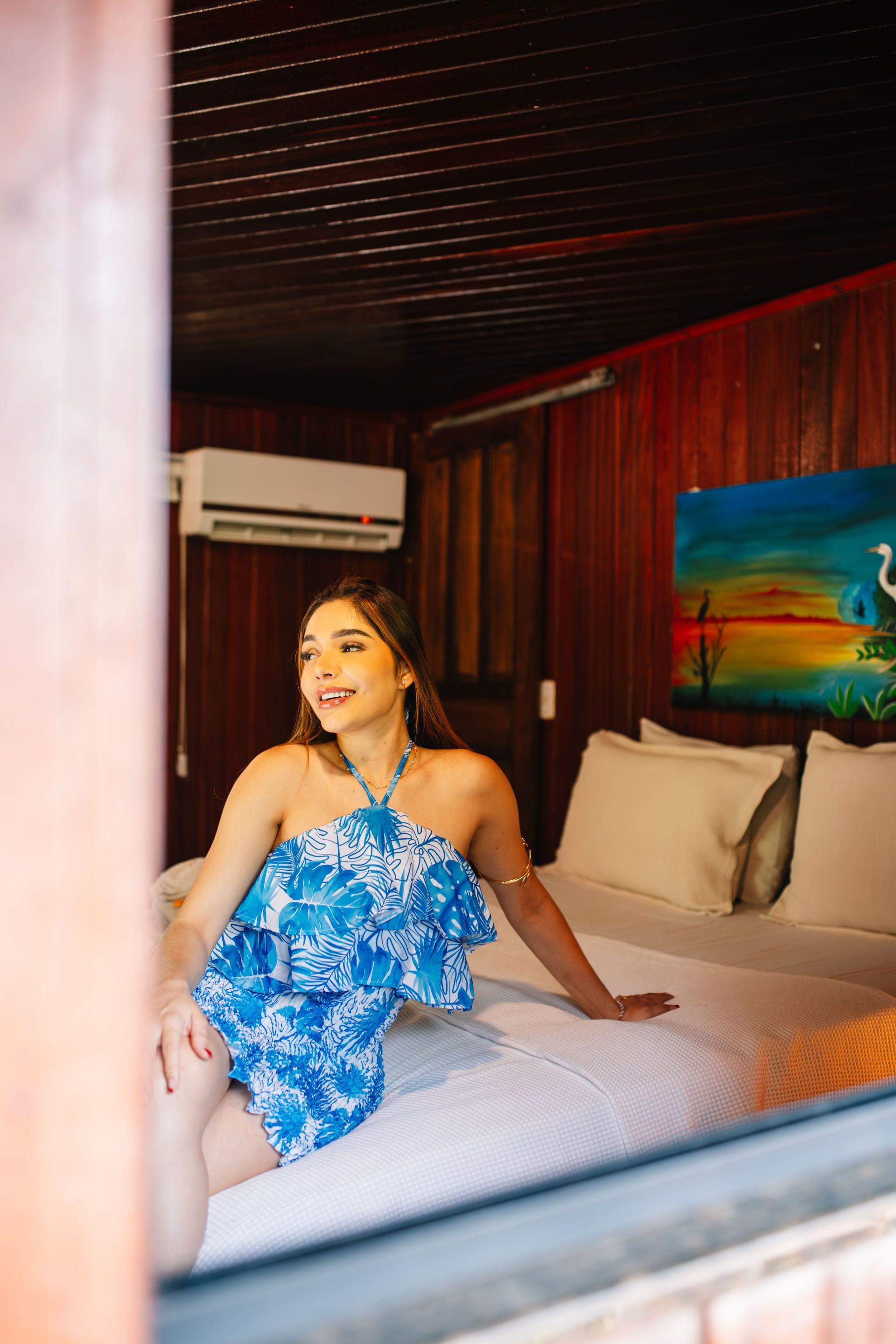 Woman in blue dress smiles on a bed in a cabin, looking towards the light.