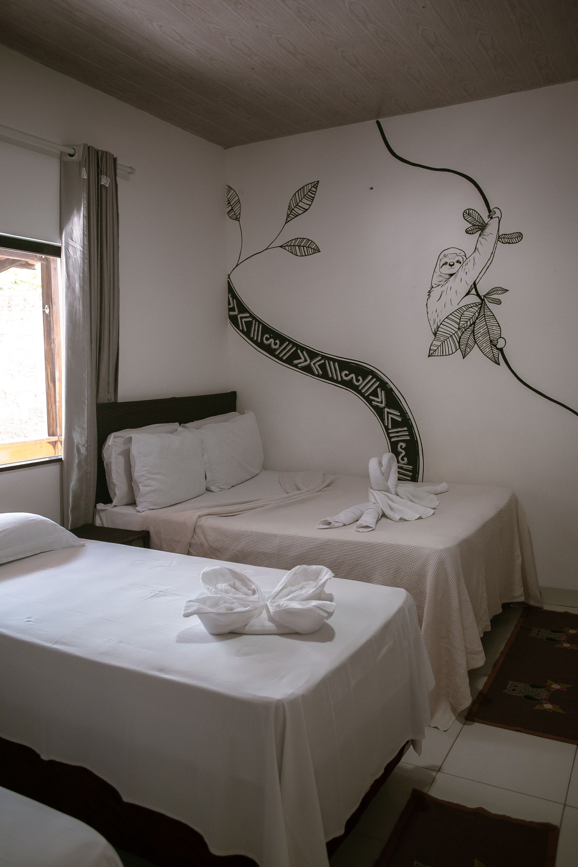 Hotel bedroom with three beds, white linens, decorative tree mural, and window with curtain.