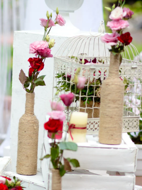 Wedding decorations: rope-wrapped bottles with roses, birdcage, candle, and white boxes, outdoors.