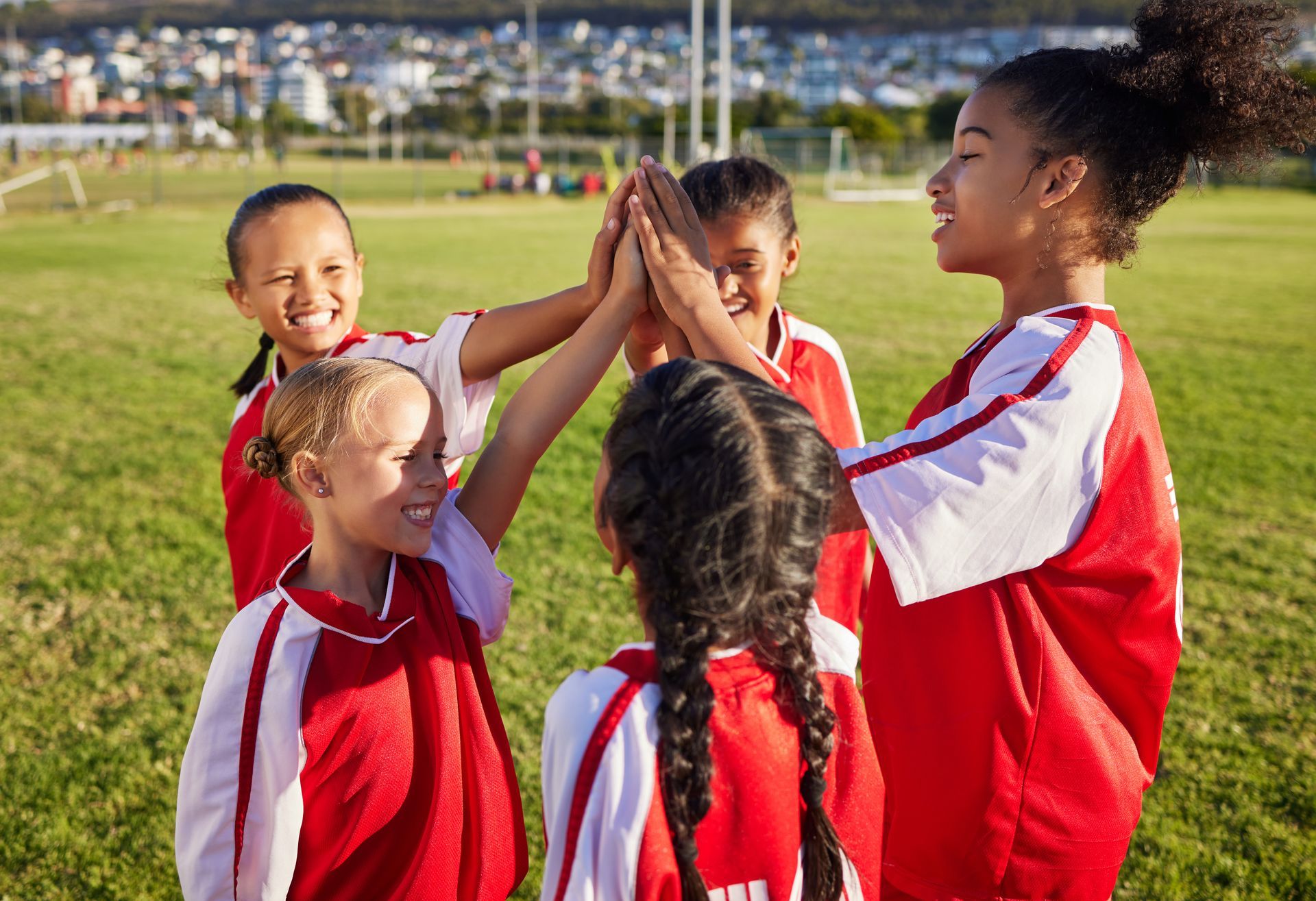 Children in red jerseys high-five on a grassy sports field on a sunny day