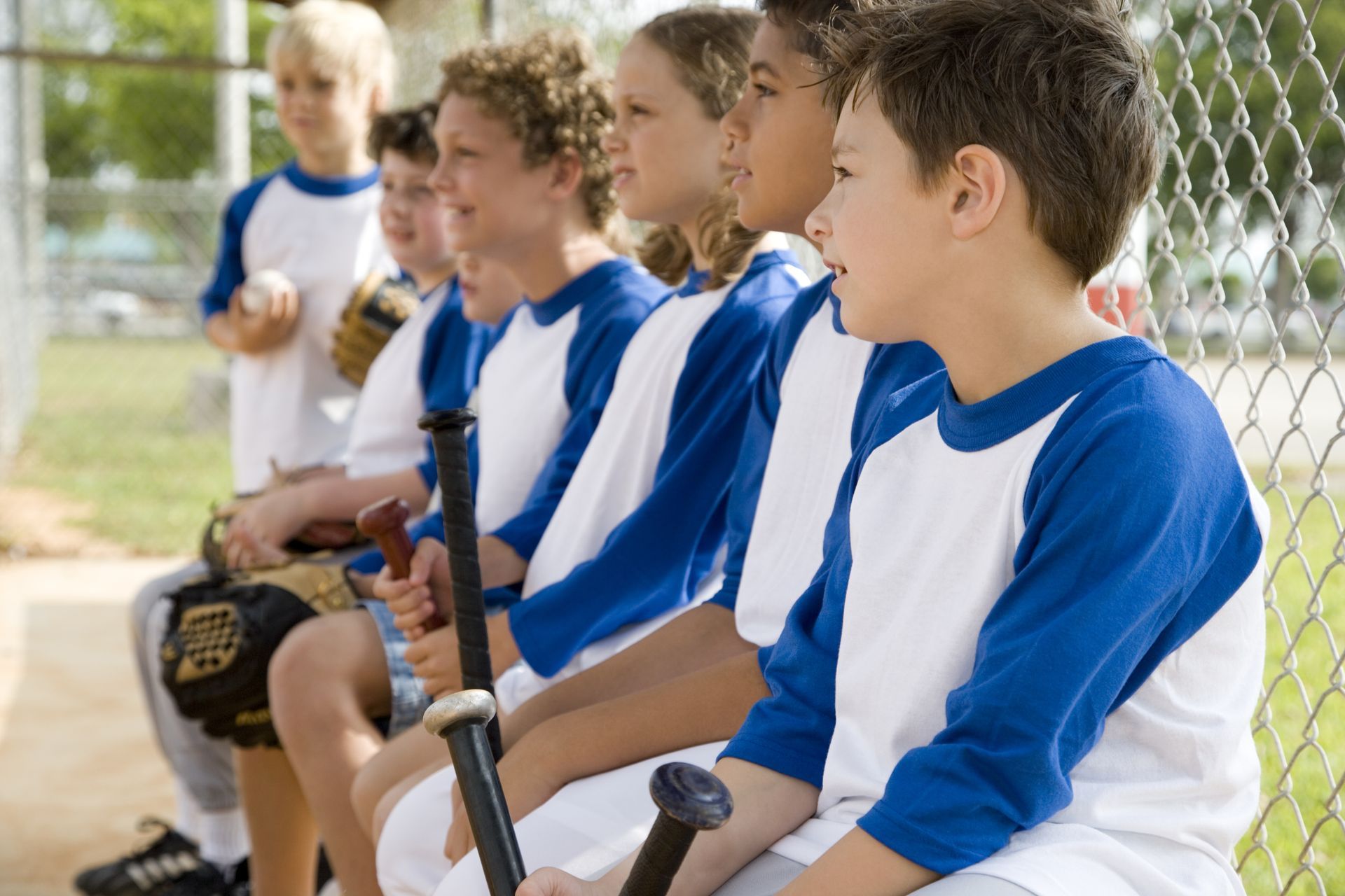 Youth baseball players in blue-and-white jerseys sit on a bench with bats and gloves, waiting on the sidelines