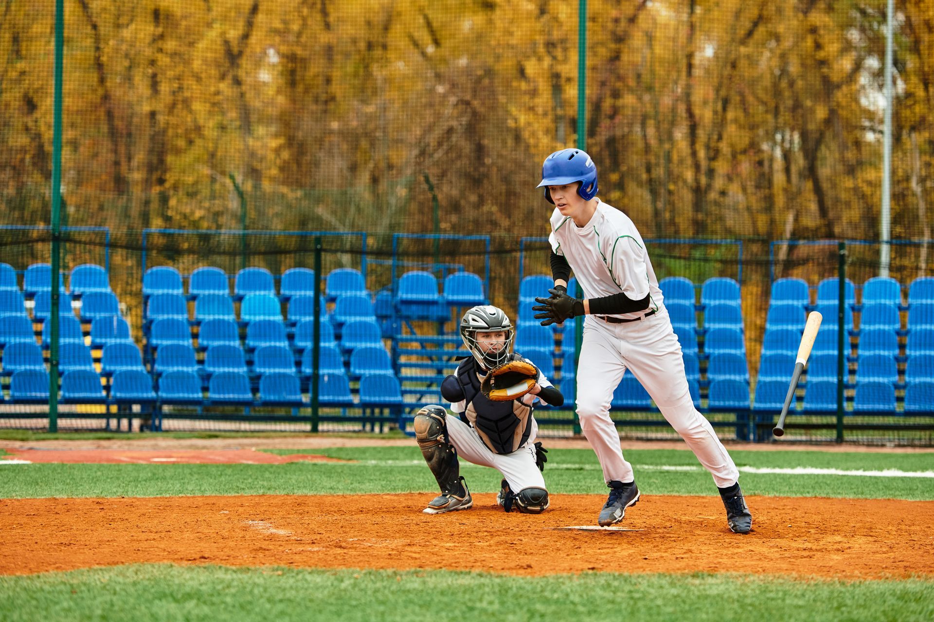 Baseball batter at the plate with catcher crouched behind, blue bleachers and autumn trees in the background