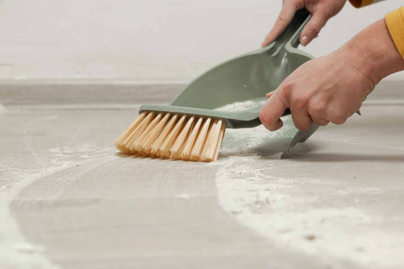 Hands sweep white dust into a dustpan on a gray floor.