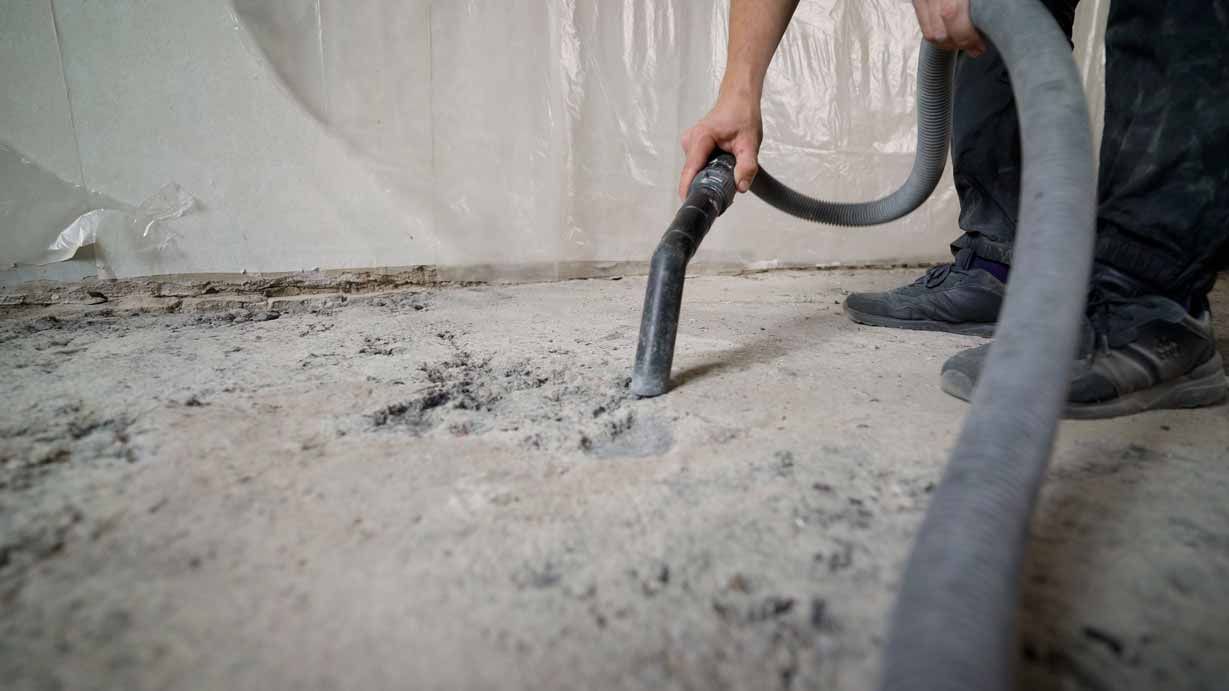 Worker using a vacuum hose to clean dust from a concrete floor indoors