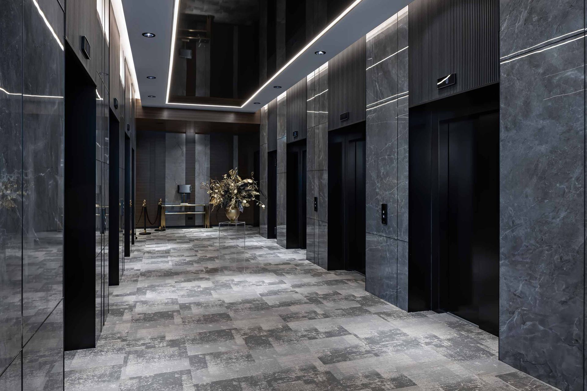 Modern hotel hallway with marble walls, dark elevator doors, and patterned carpet leading to floral arrangement