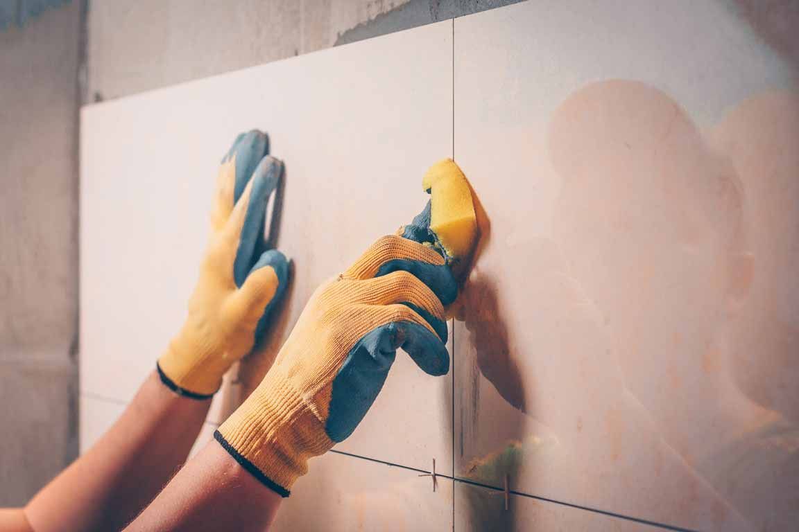 Hands wearing yellow gloves install large white wall tiles with adhesive sponge.