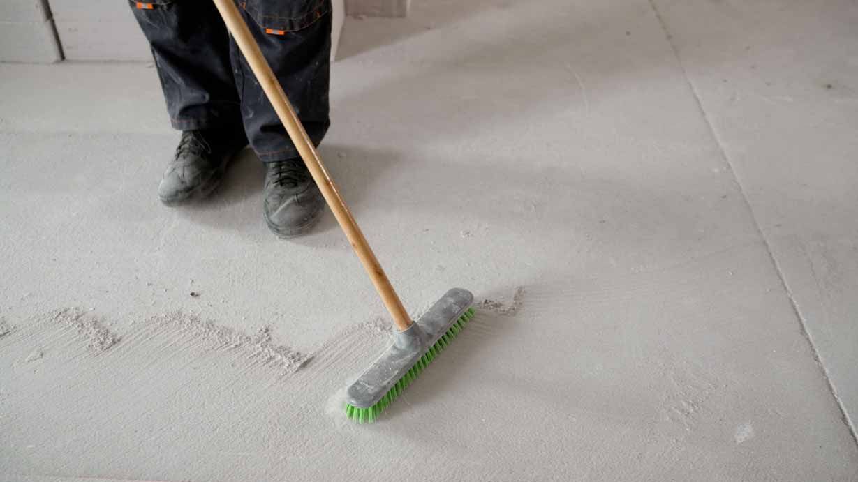 Person sweeping dust on a concrete floor with a broom and green brush head.