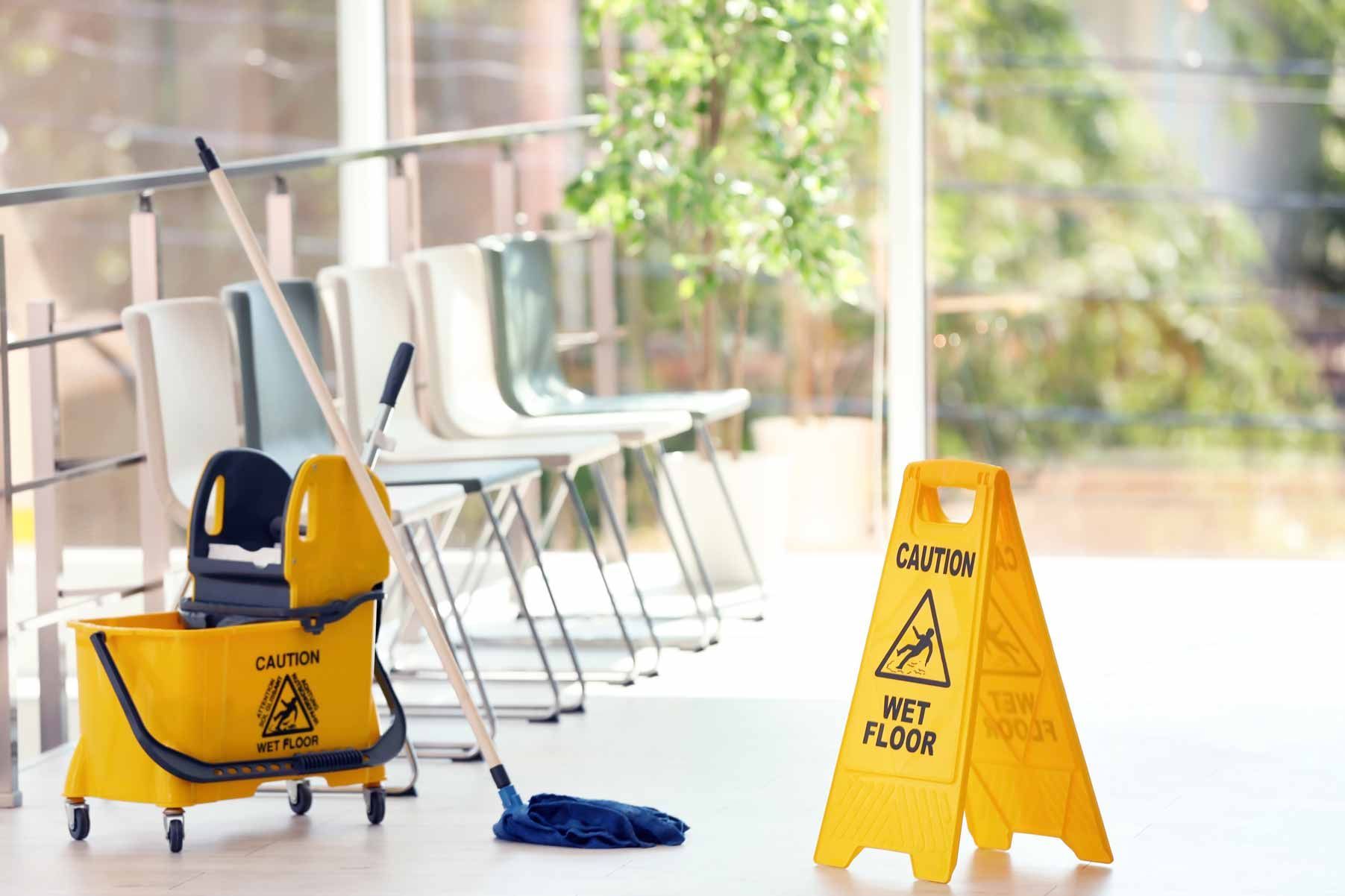 Yellow wet-floor sign and mop in a bright hallway with stacked chairs and a cleaning cart.
