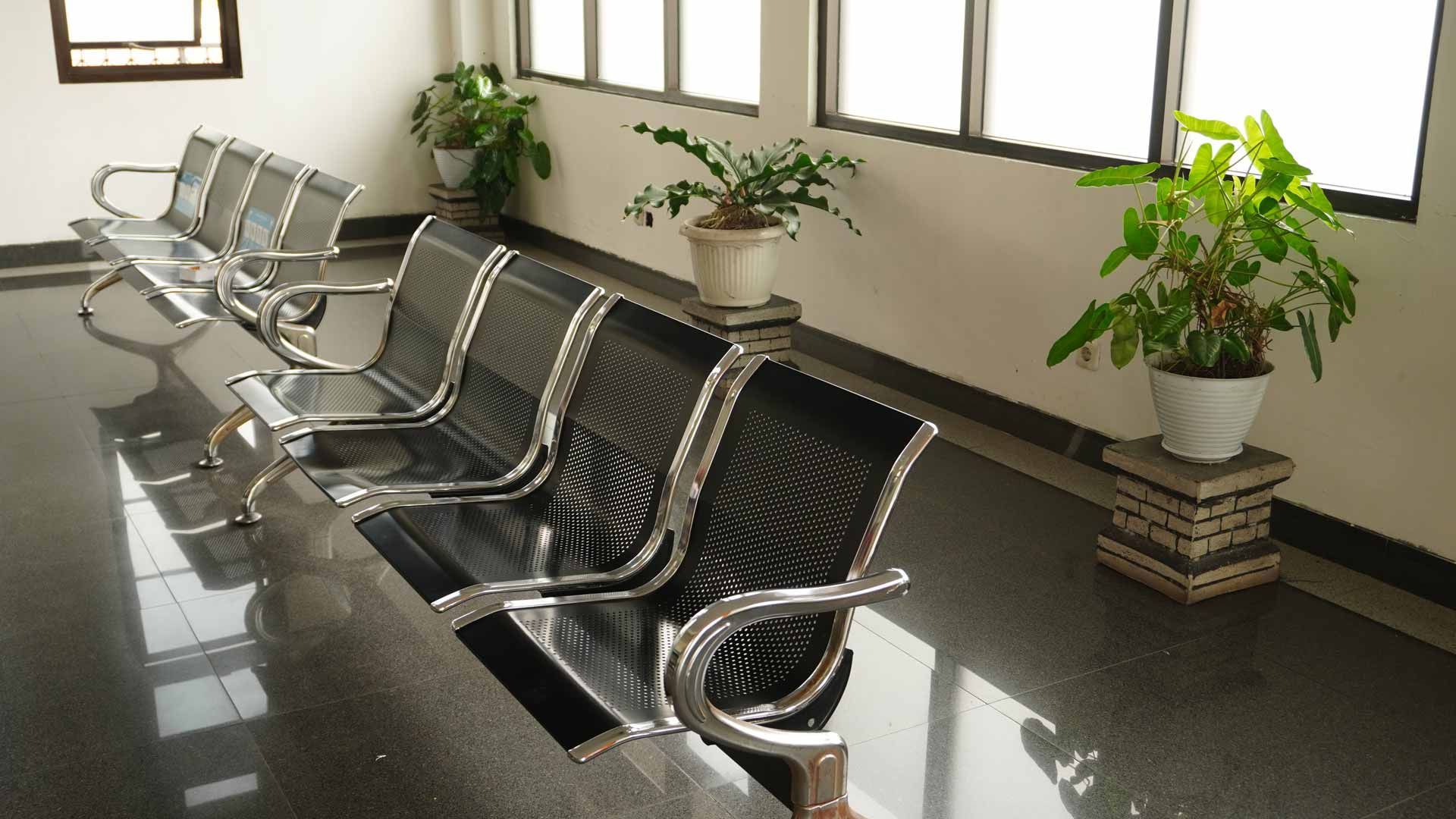 Empty waiting area with black metal benches, potted plants, and glossy tiled floor beside windows