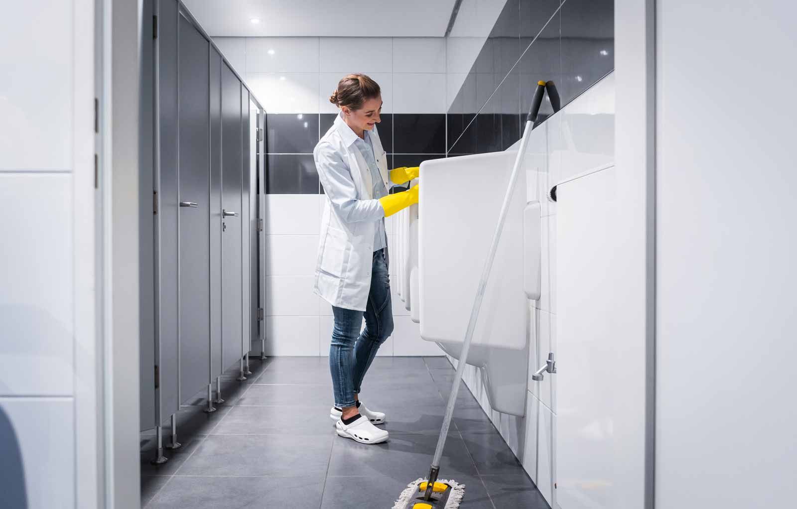 Cleaner in a lab corridor wiping a white cabinet with yellow gloves and a mop nearby