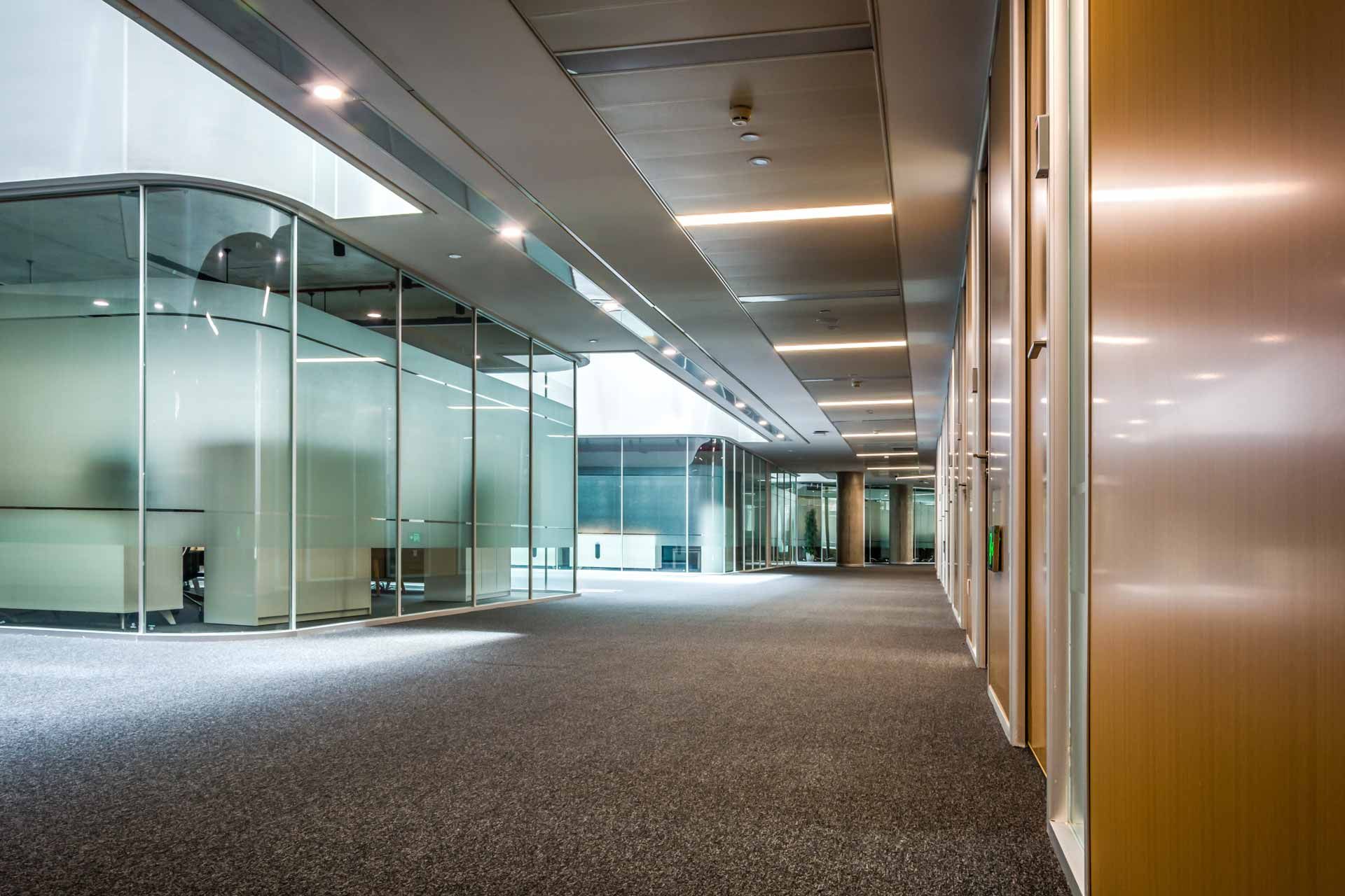 Modern office hallway with glass-walled rooms, carpeted floor, and metallic doors