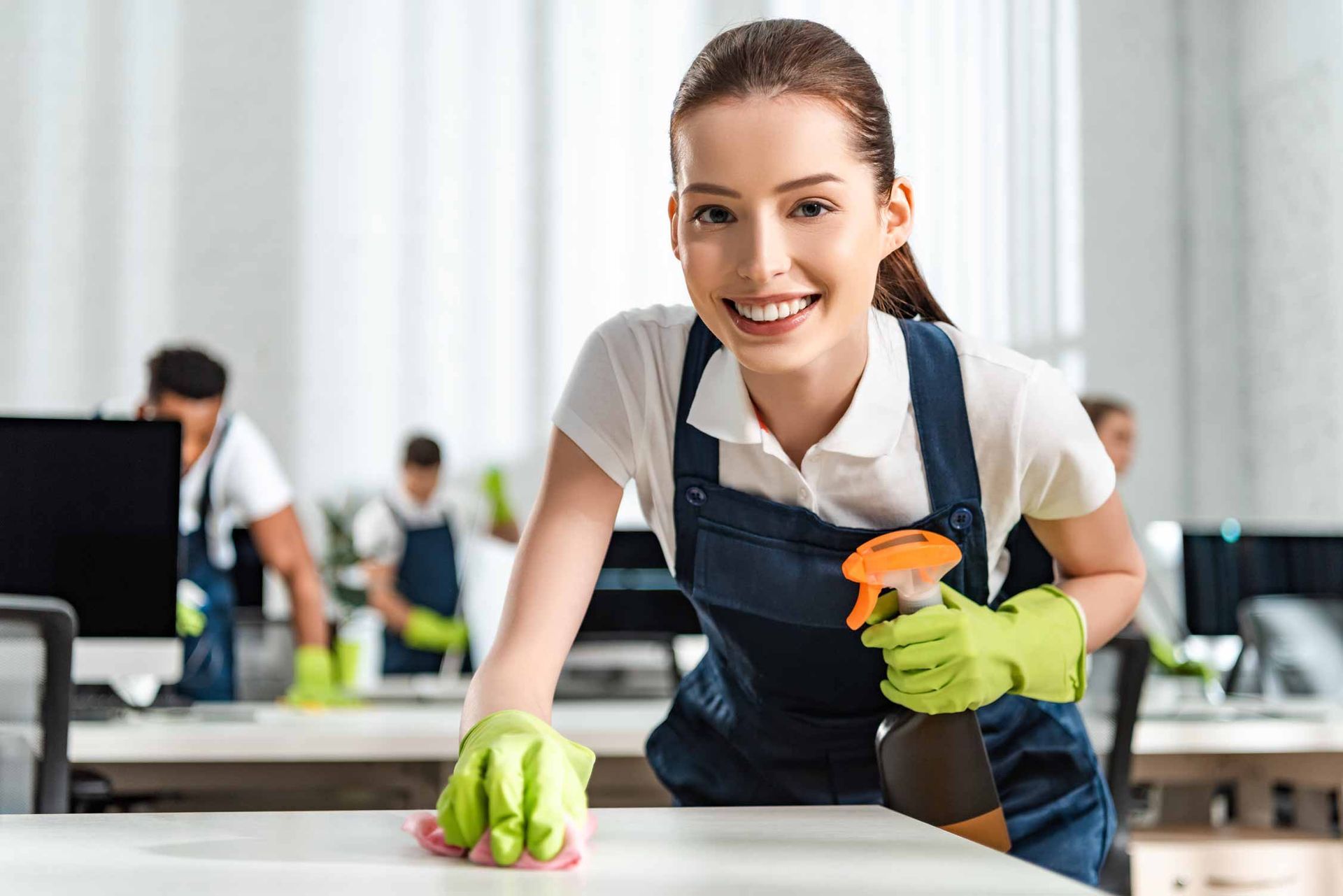 Smiling cleaner in apron wiping a desk with a cloth and spray bottle in an office.