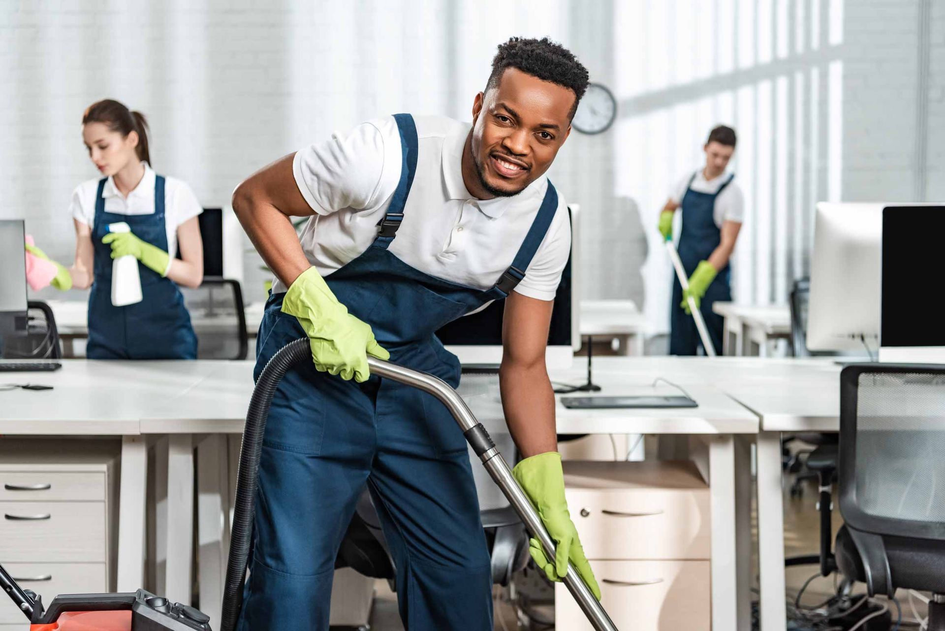Office cleaner vacuuming a desk area with coworkers in the background