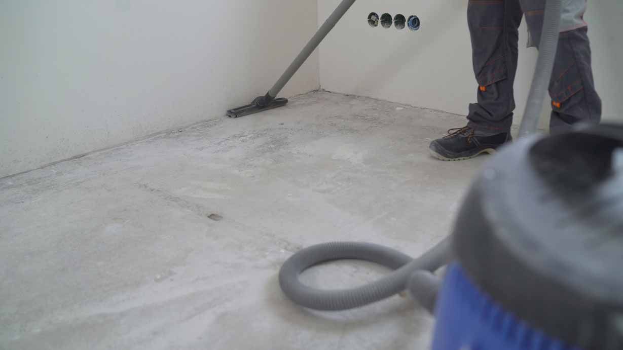 Person vacuuming a dusty room corner with a blue vacuum cleaner and hose