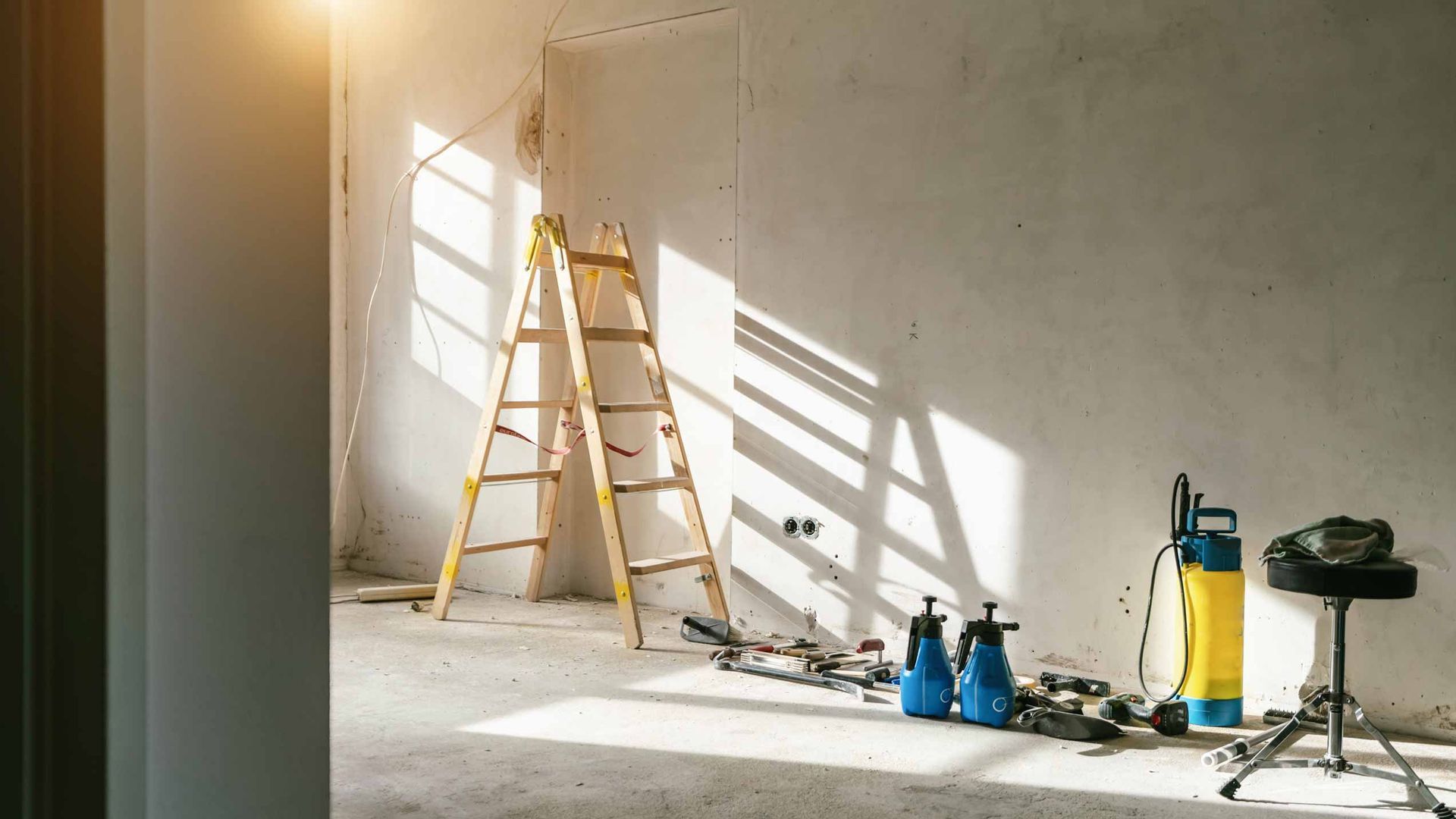 Empty room under renovation with ladder, tools, paint cans, and a work light by a sunlit wall
