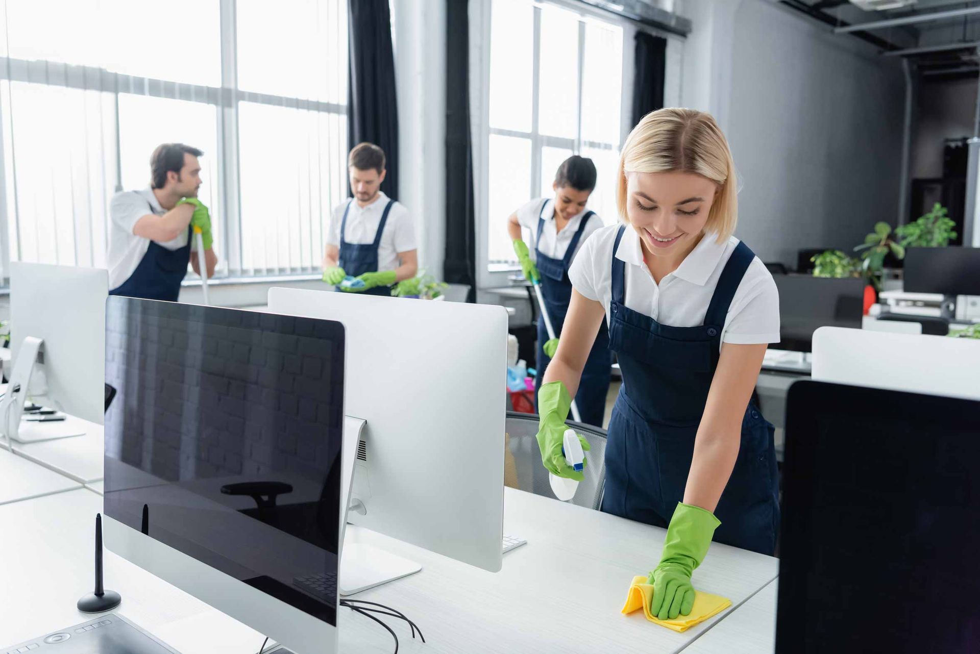 Office workers cleaning desks in a bright open-plan office, with monitors and green gloves.