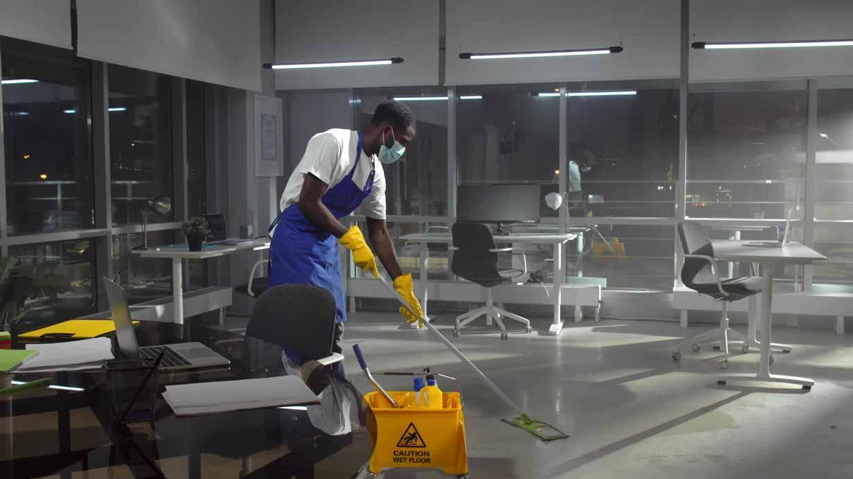 Janitor mops a wet floor in a modern office break room, with a yellow caution sign nearby.