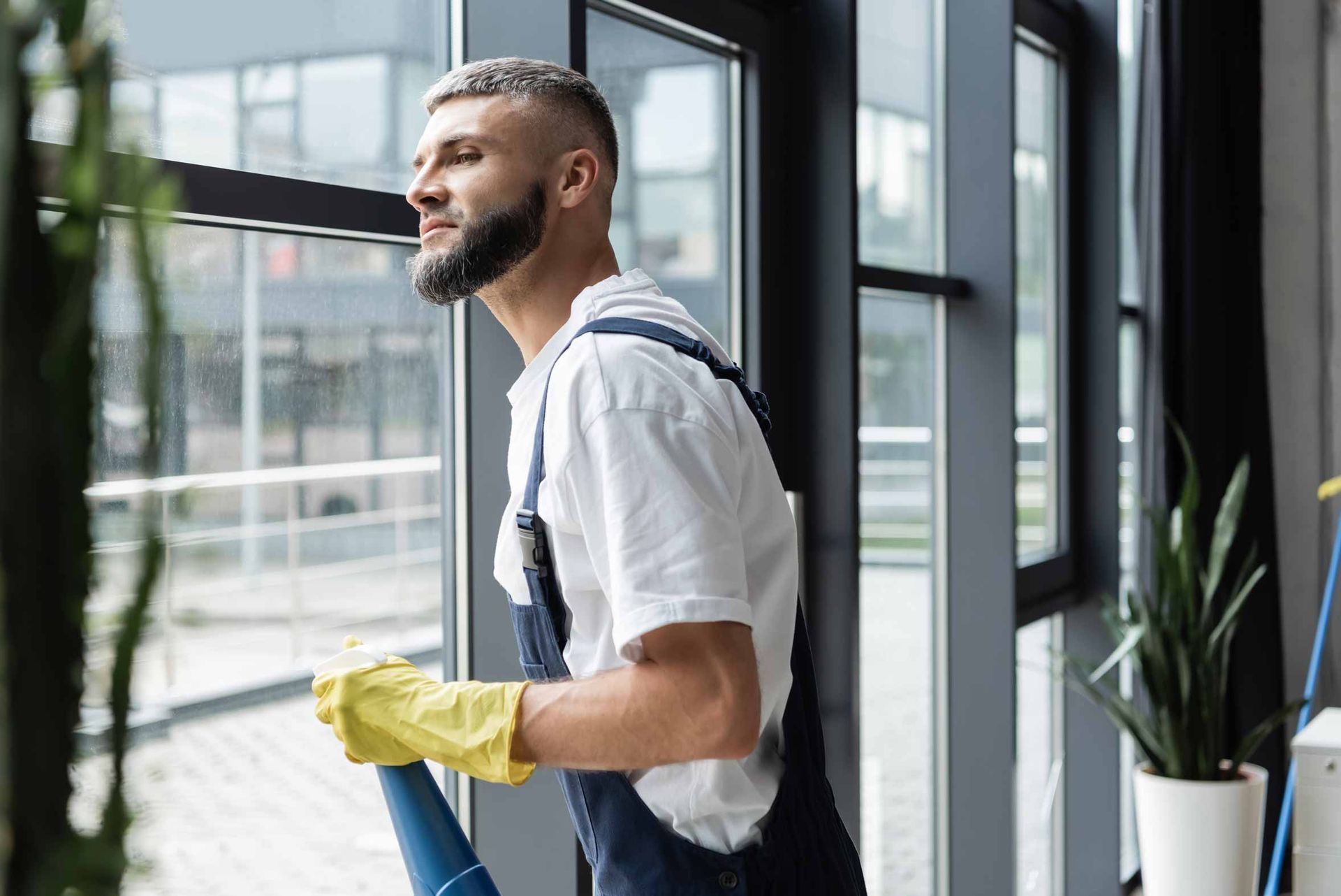 Window cleaner in a bright office, wearing yellow gloves and carrying a cleaning tool.