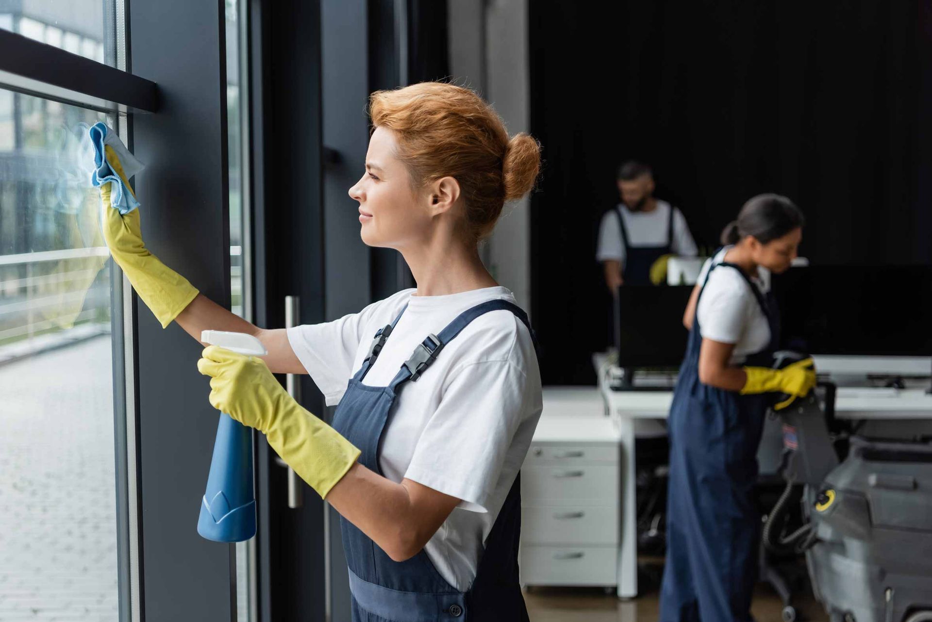 Two cleaners in aprons and yellow gloves cleaning windows in an office workspace