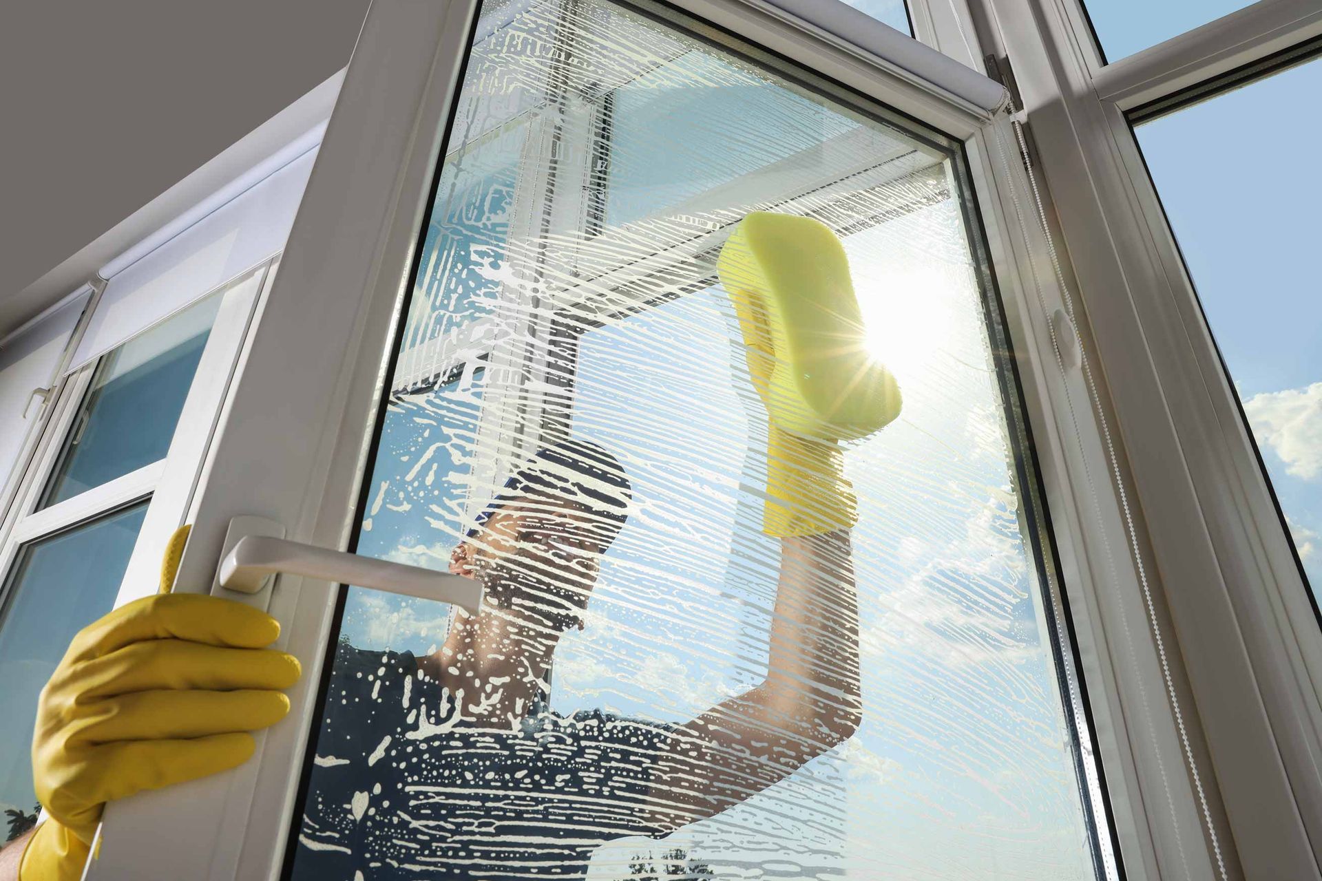 Person cleaning a glass door with yellow gloves and a sponge, seen through sunlight.