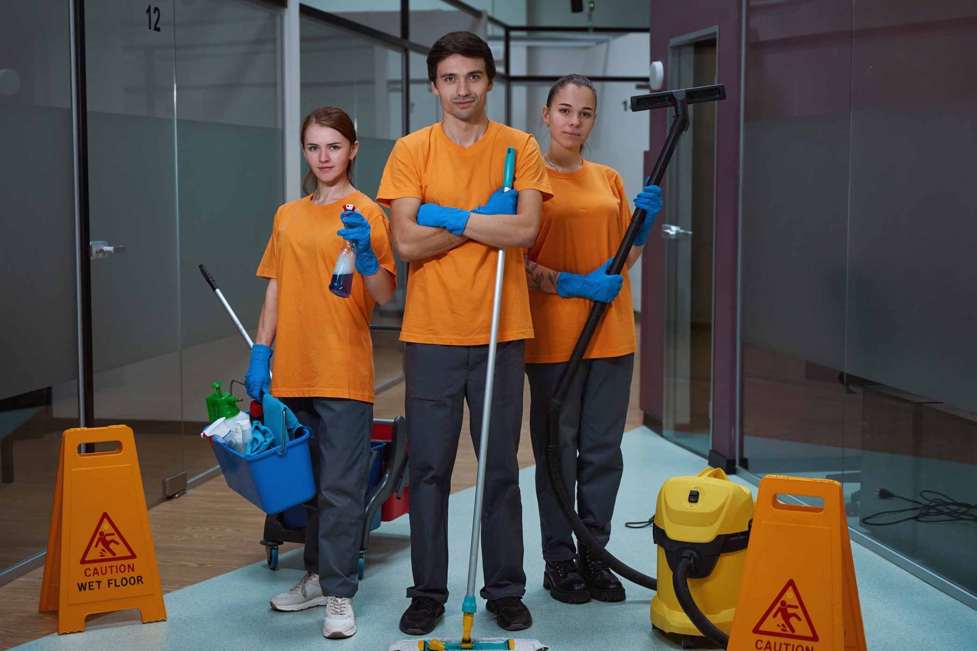 Three cleaners in orange uniforms holding mops and supplies in a lobby near wet-floor signs