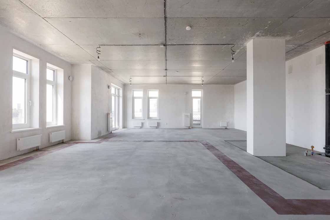 Empty unfinished room with concrete floor, white walls, large windows, and exposed ceiling fixtures.