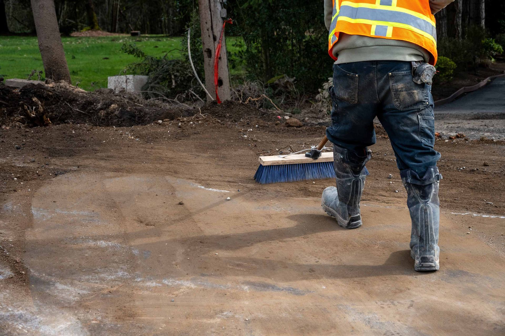 Worker in a high-visibility vest and rubber boots sweeping muddy ground at a construction site