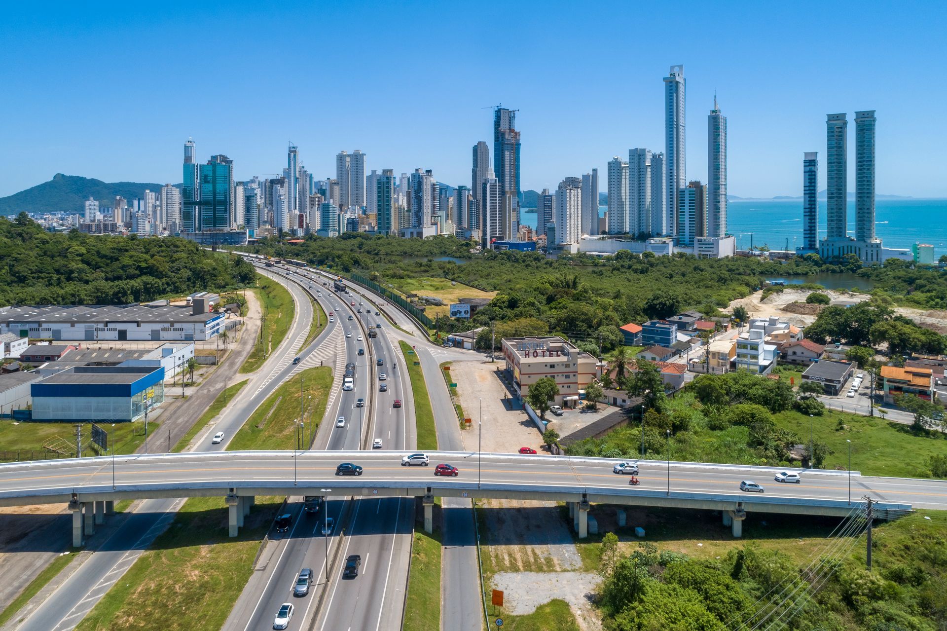 Vista aérea mostra a rodovia movimentada e um viaduto em primeiro plano, com a paisagem urbana de BC