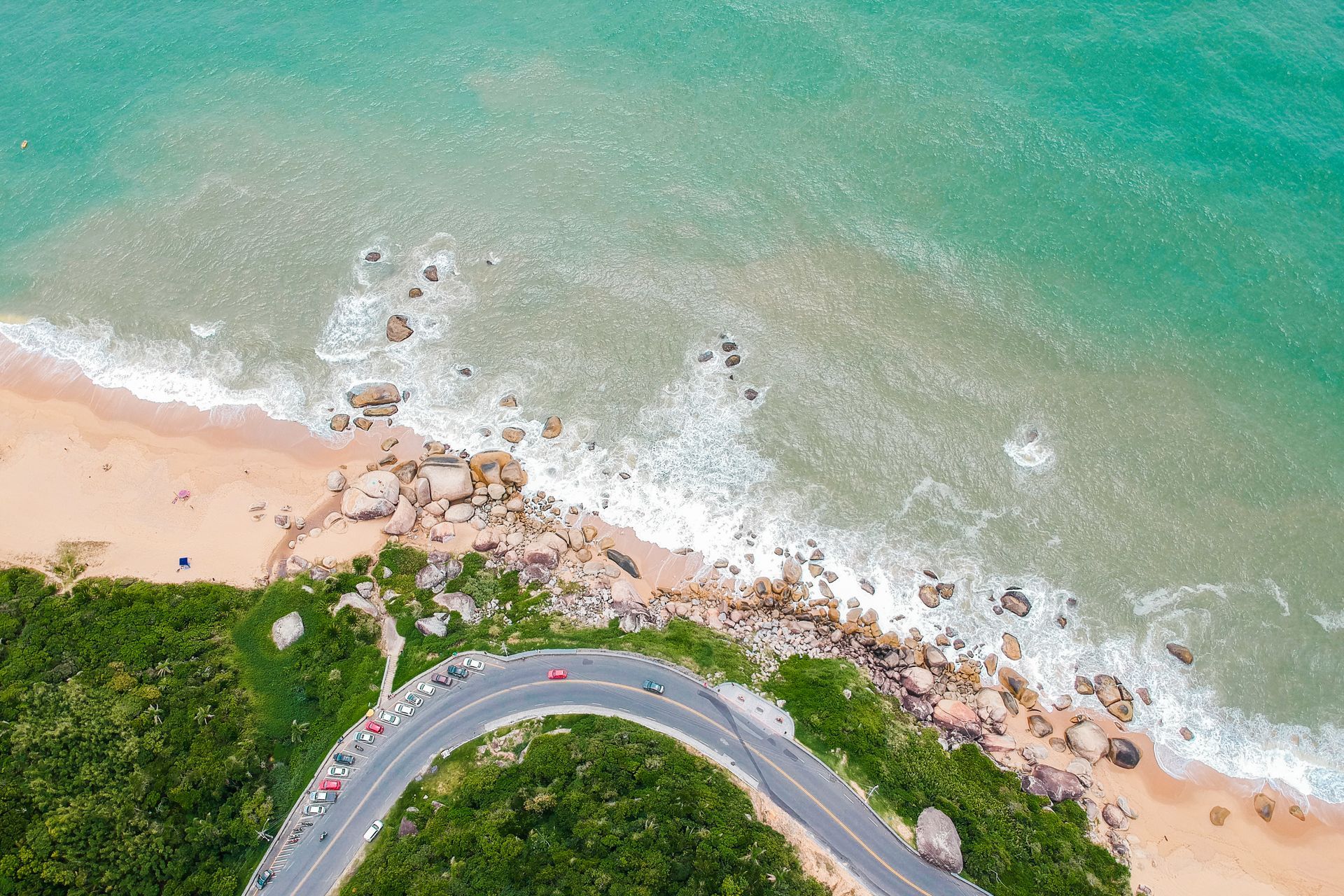 Vista aérea de uma estrada litorânea cercada por vegetação, areia e o mar. 